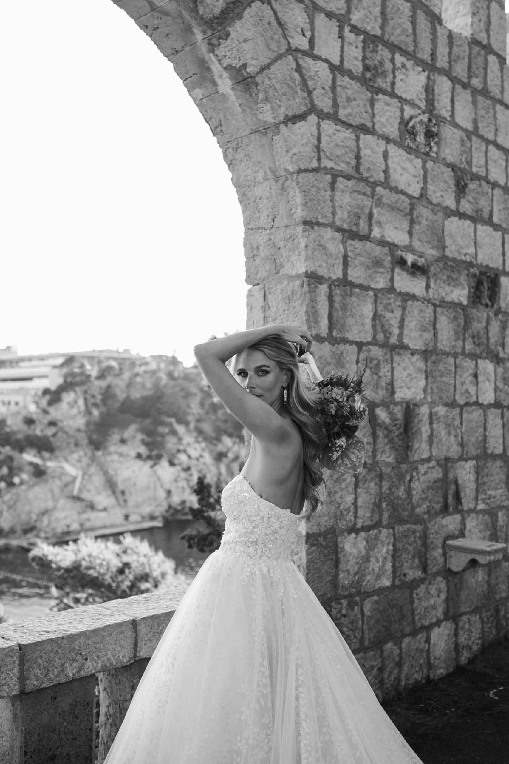 Elegantna crno-bijela fotografija mladenke u raskošnoj vjenčanici. Pozira na kamenoj terasi pod lukom, držeći ruke uz kosu, s pogledom na dubrovačku obalu u pozadini.