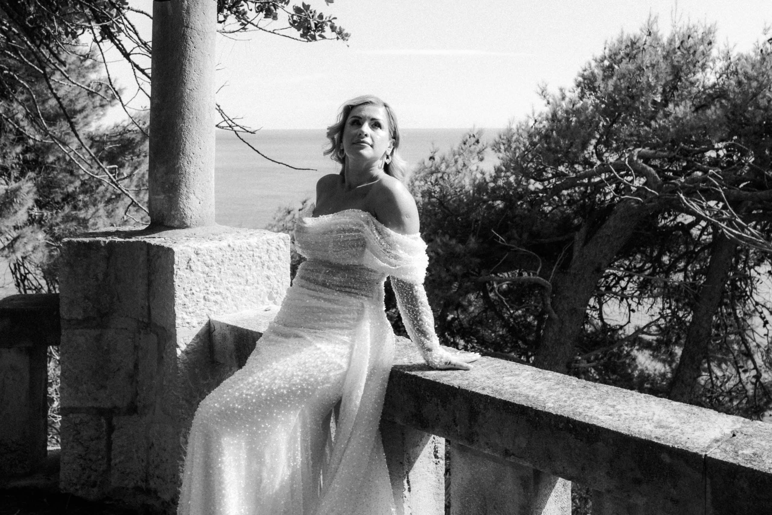 legantna mladenka u raskošnoj vjenčanici sjedi na kamenoj ogradi balkona okružena mediteranskim borovima. Bezvremenska filmska crno-bijela fotografija.