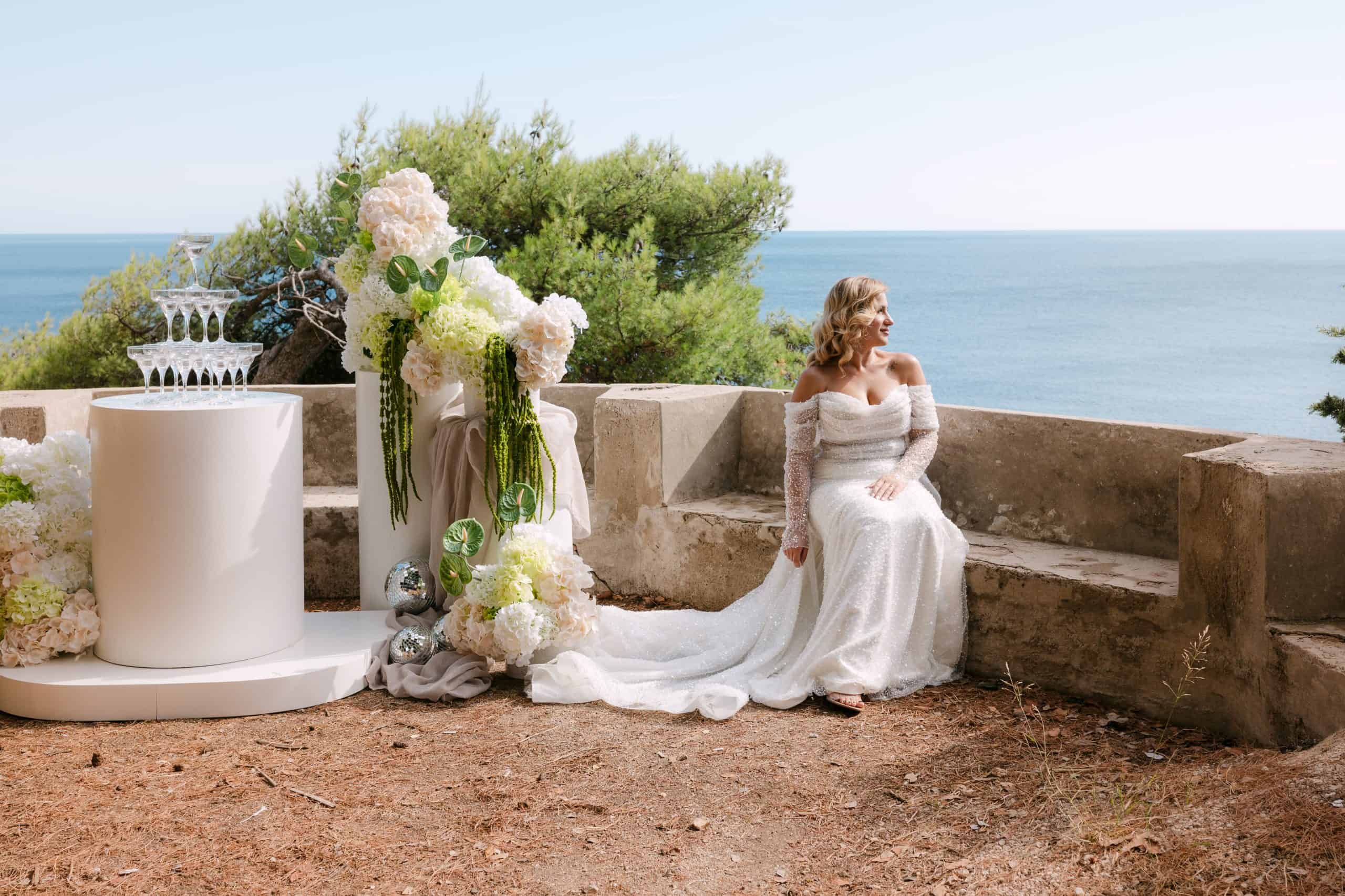 Prekrasna mladenka u raskošnoj vjenčanici s dugačkim šlepom sjedi na kamenoj ogradi pored svadbenog oltara s cvijećem i kulom od šampanjca.