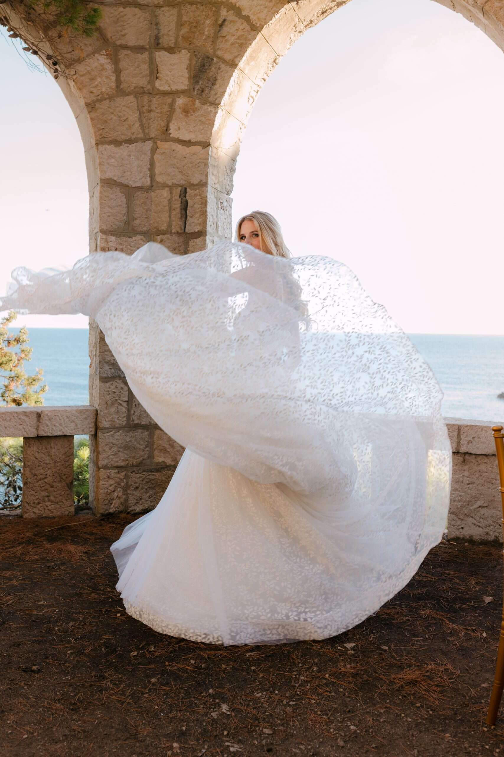 Editorial fotografija mladenke u bijeloj svjetlucavoj vjenčanici. Mladenka elegantno maše svojim velom u zraku pozirajući ispod kamenog luka okrenuta prema Jadranskom moru.