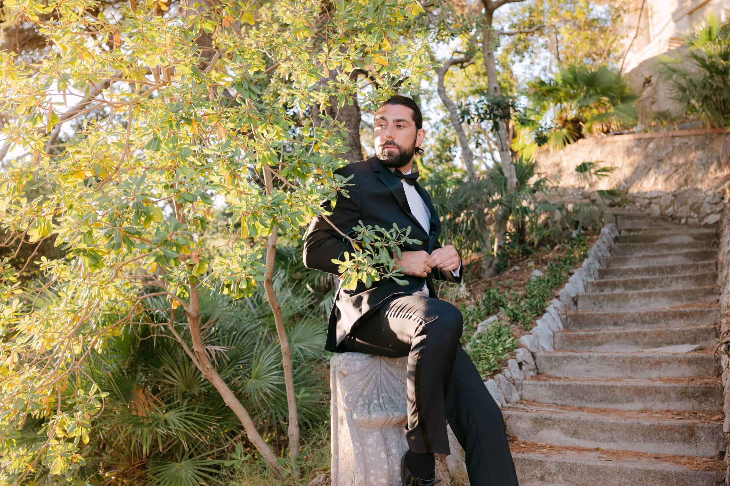 Editorial portret mladoženje u elegantnom crnom odijelu. Zamišljeno sjedi na starom kamenom zidiću pored povijesnih stepenica okružen bujnim mediteranskim drvećem u Dubrovniku.