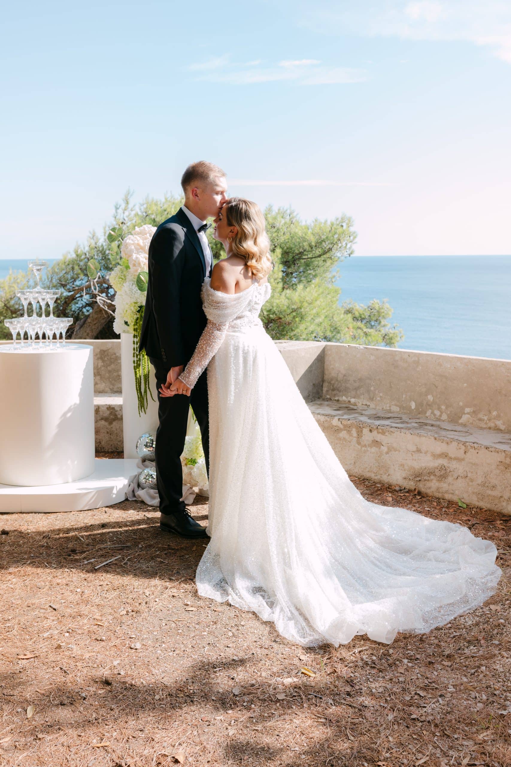 Široki kadar mladenaca koji stoje uz kameni zid s predivnim pogledom na more. Mladoženja ljubi mladenku pored elegantne kule od čaša za šampanjac na terasi.