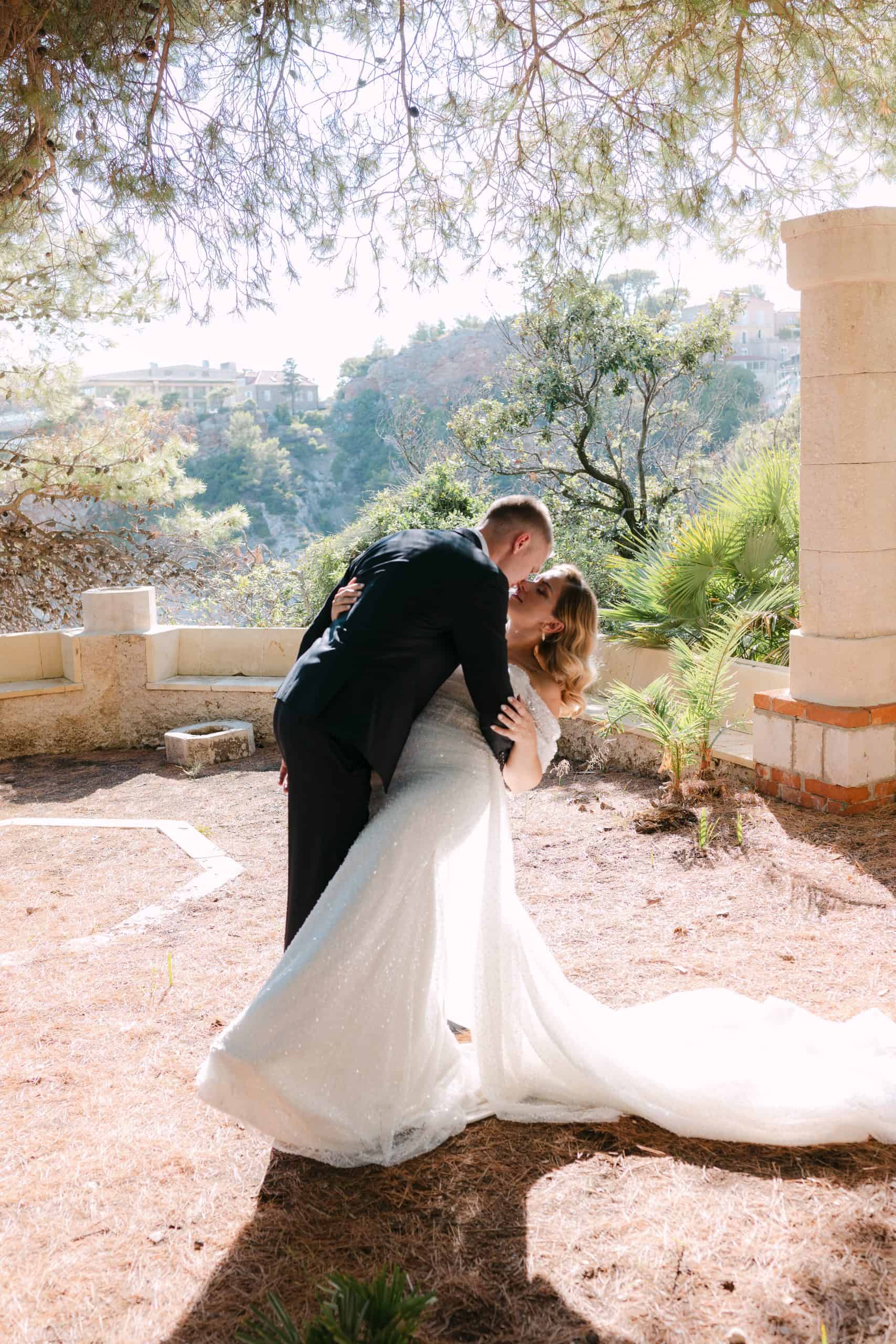 Mladoženja drži mladenku u romantičnom nagibu i ljubi je ispred kamenog zida s pogledom na mediteranski krajolik. Wedding session obasjan suncem u Vili Čingrija.