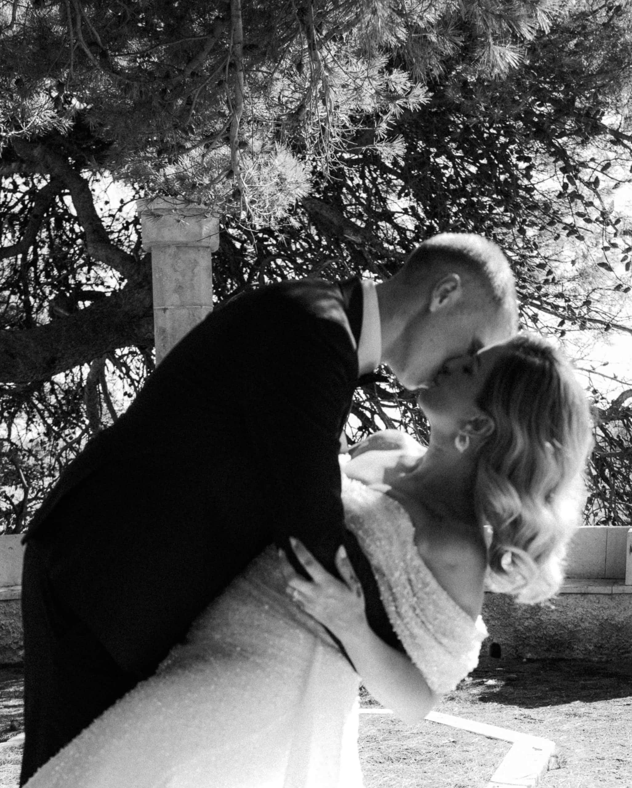 Crno-bijela fotografija mladenaca. Mladoženja drži mladenku u naručju i ljubi je ispred starog kamenog stupa okružen drvećem u Dubrovniku.