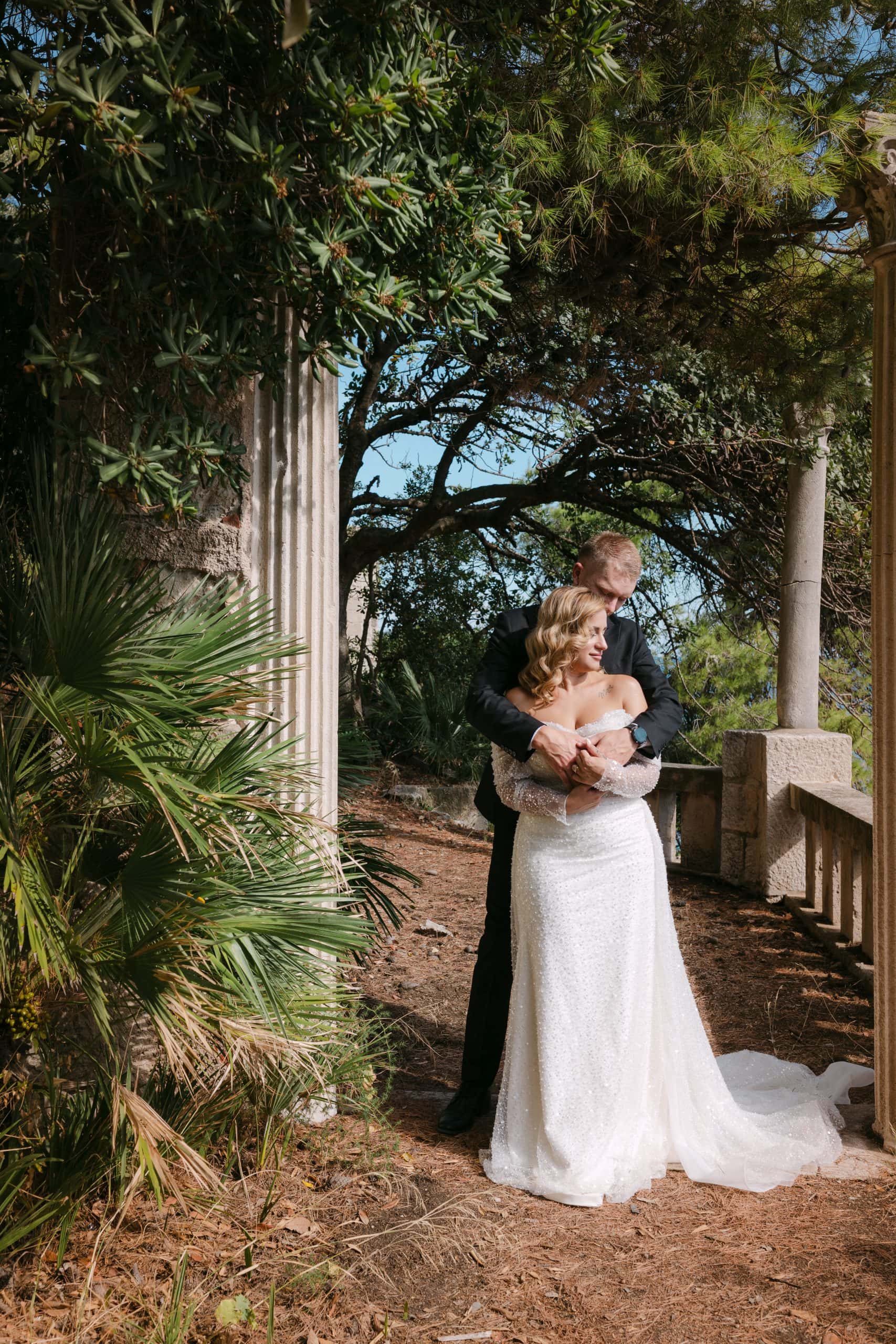 Mladoženja s leđa nježno grli mladenku dok stoje u sjenovitom mediteranskom vrtu pored kamenog stupa i palmi u Vili Čingrija.