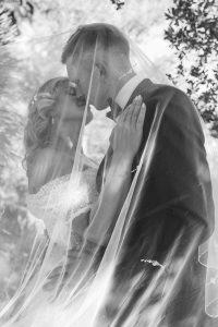 Crno-bijela svadbena fotografija mladenaca u zagrljaju ispod vela. Bezvremenski i romantičan kadar s naglašenim filmskim zrnom.