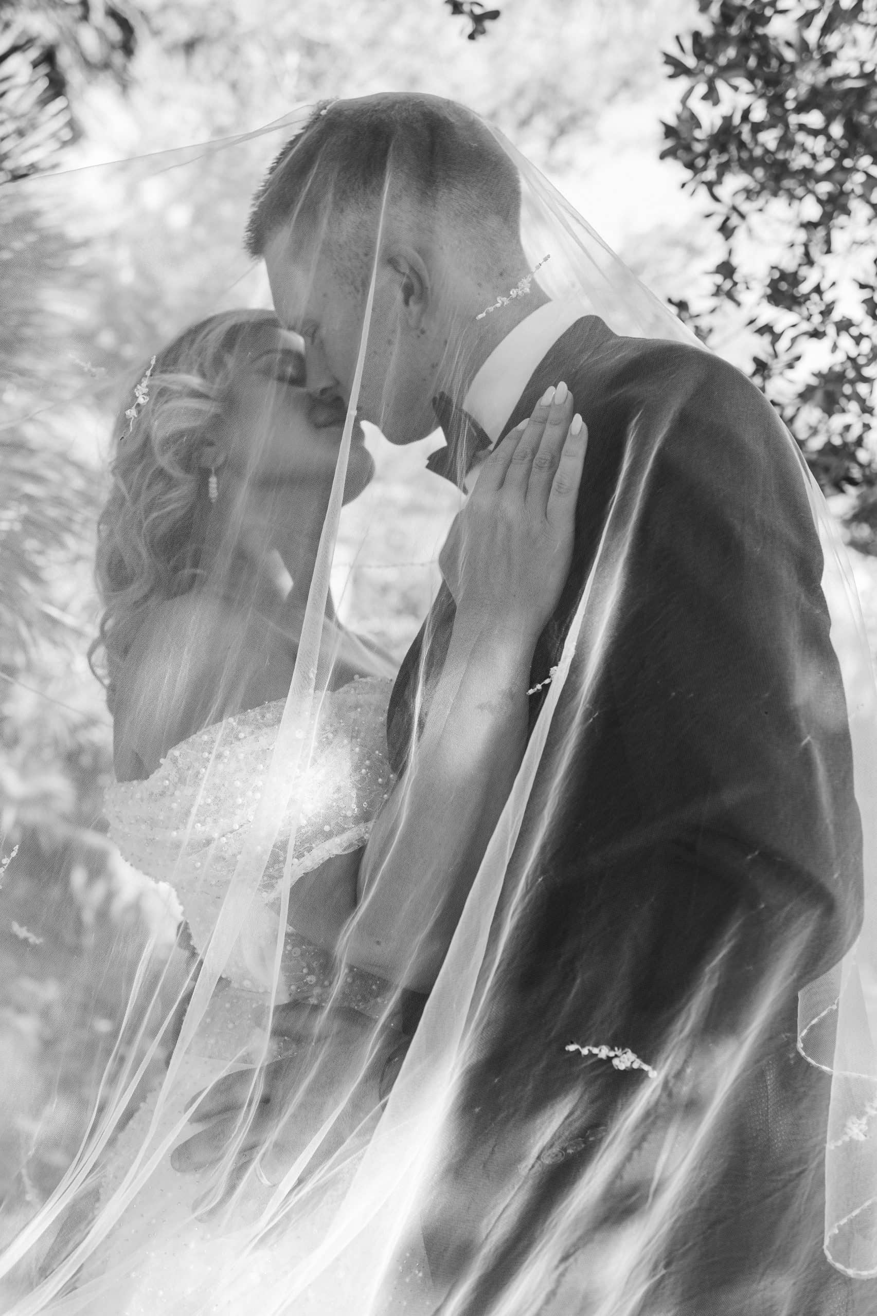 Crno-bijela svadbena fotografija mladenaca u zagrljaju ispod vela. Bezvremenski i romantičan kadar s naglašenim filmskim zrnom.