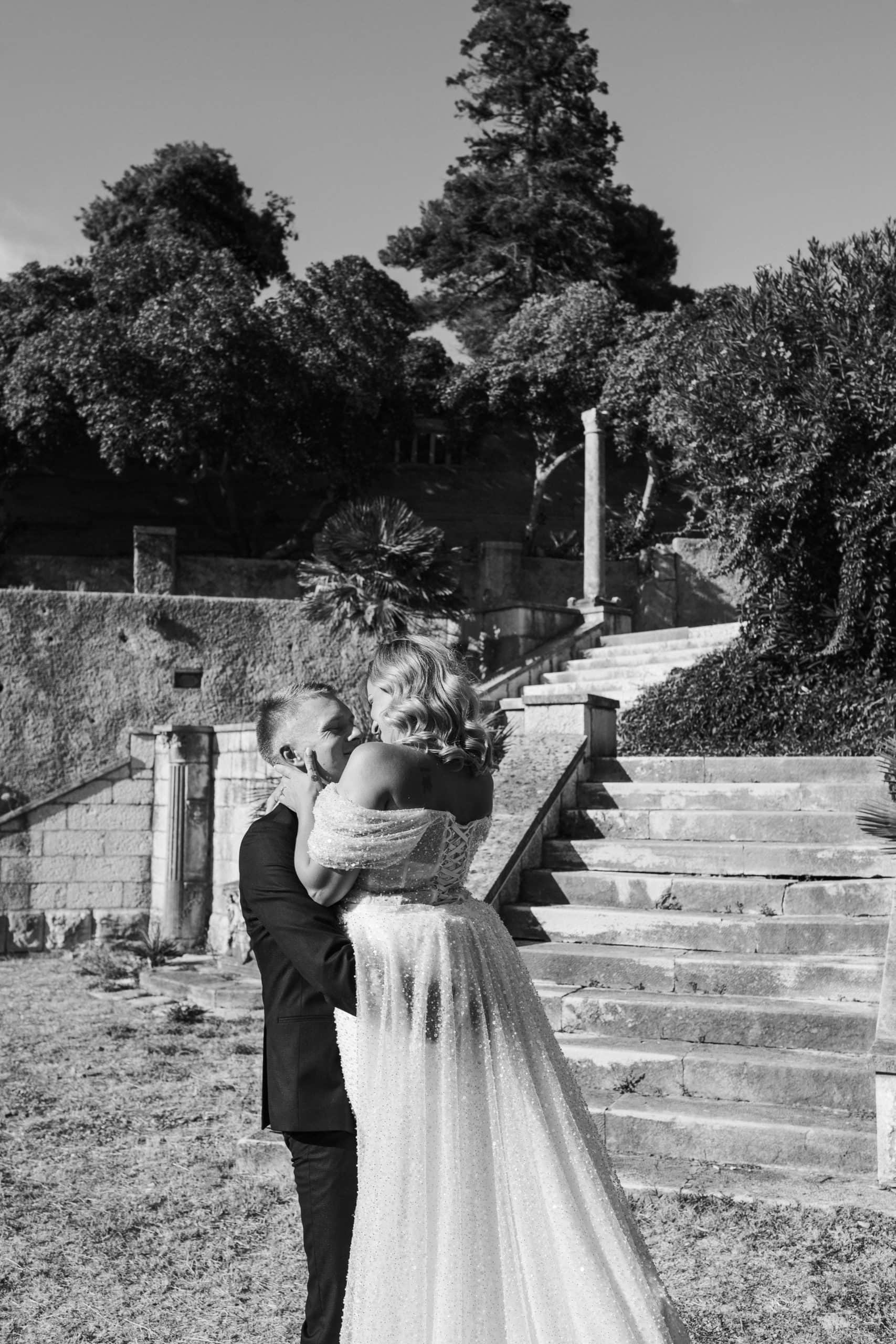 Crno-bijela svadbena fotografija mladoženje koji drži mladenku ispred povijesnih kamenih stepenica Vile Čingrija u Dubrovniku.