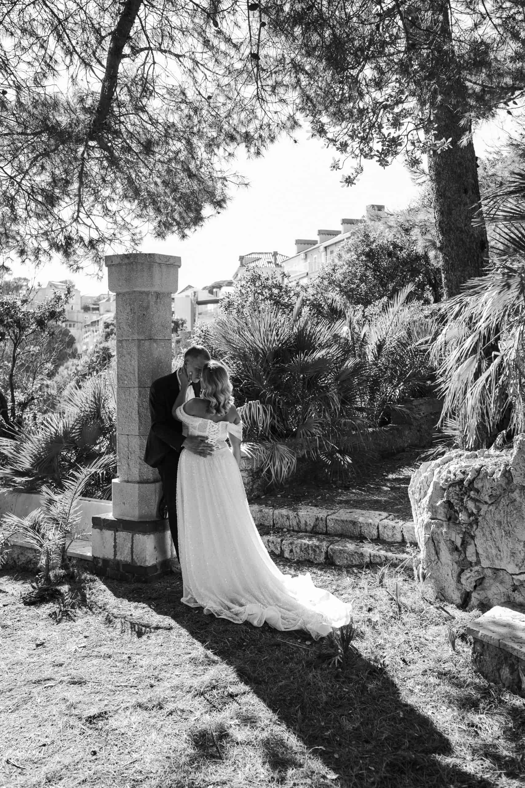 Crno-bijela fotografija mladenaca u zagrljaju ispod povijesnog kamenog stupa i drveća u Vili Čingrija. Jak kontrast svjetla i sjene stvara dramatičan filmski ugođaj.