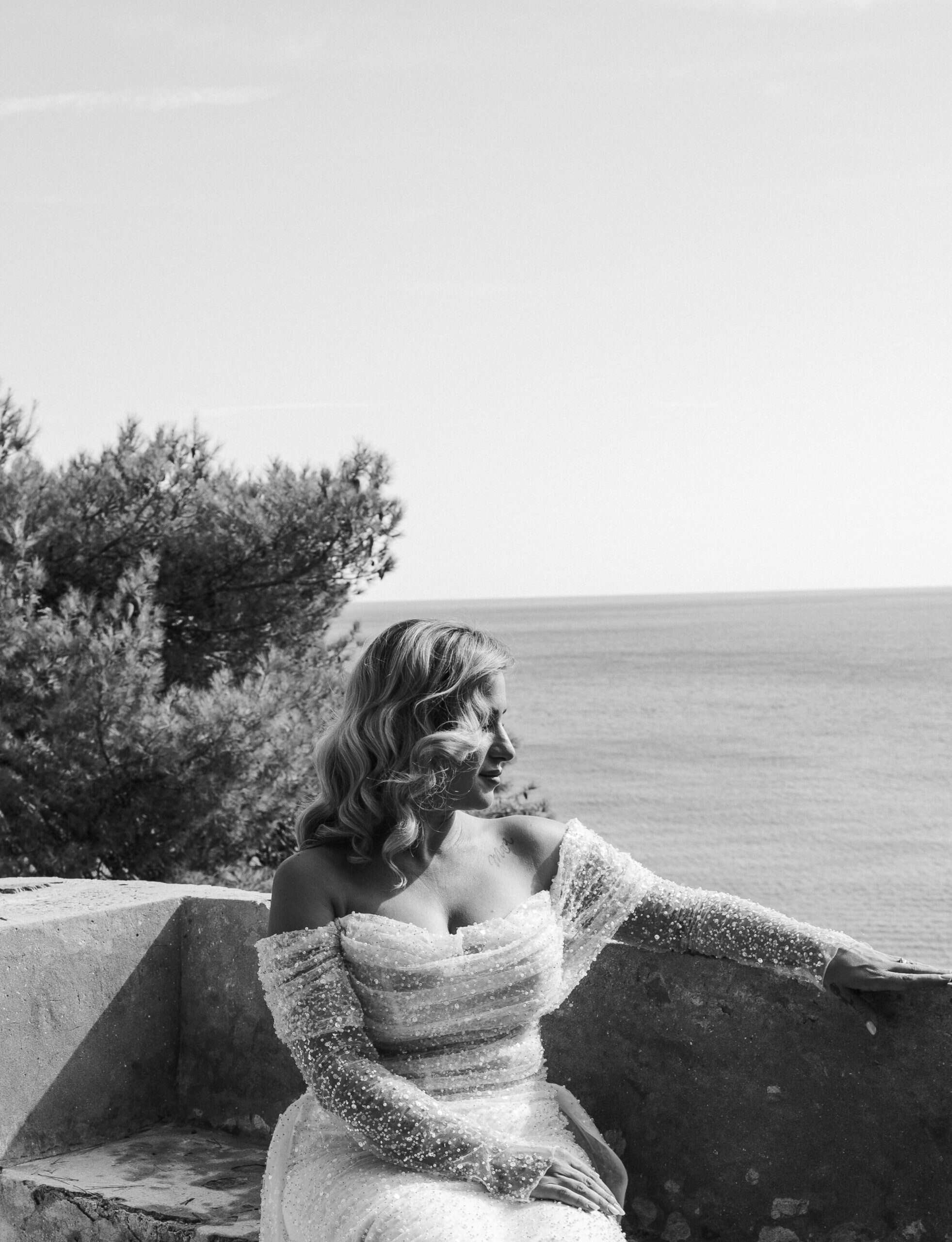 Elegantna crno-bijela fotografija mladenke koja sjedi na kamenoj ogradi obasjana suncem, s predivnim pogledom na mirni morski horizont.