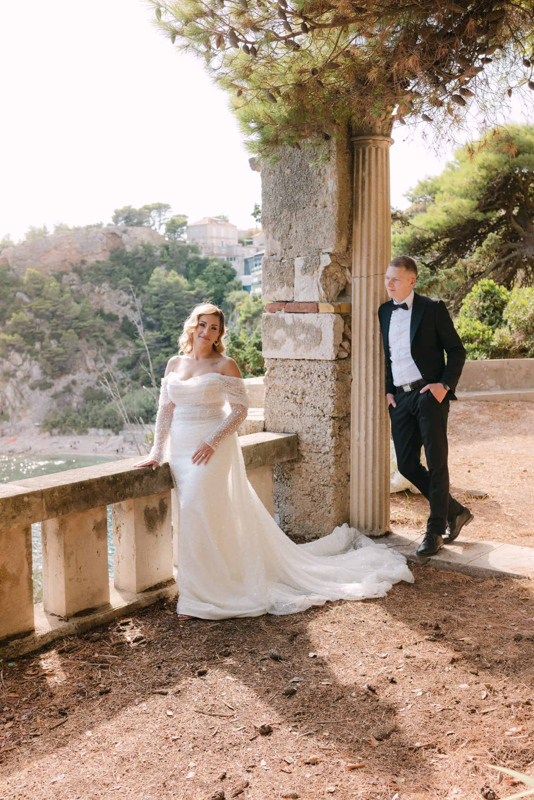 Široki kadar mladenaca na povijesnoj kamenoj terasi Vile Čingrija u Dubrovniku. Mladenka stoji uz ogradu s pogledom na uvalu, dok mladoženja u crnom odijelu stoji naslonjen na masivni kameni stup okružen borovima.