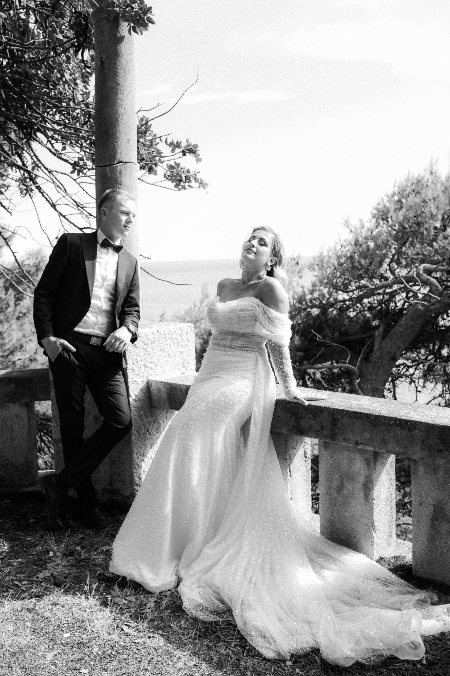 Dokumentarna crno-bijela svadbena fotografija na terasi. Mladoženja je naslonjen na kameni stup, dok mladenka sjedi na ogradi s dugim šlepom vjenčanice koji dramatično pada.