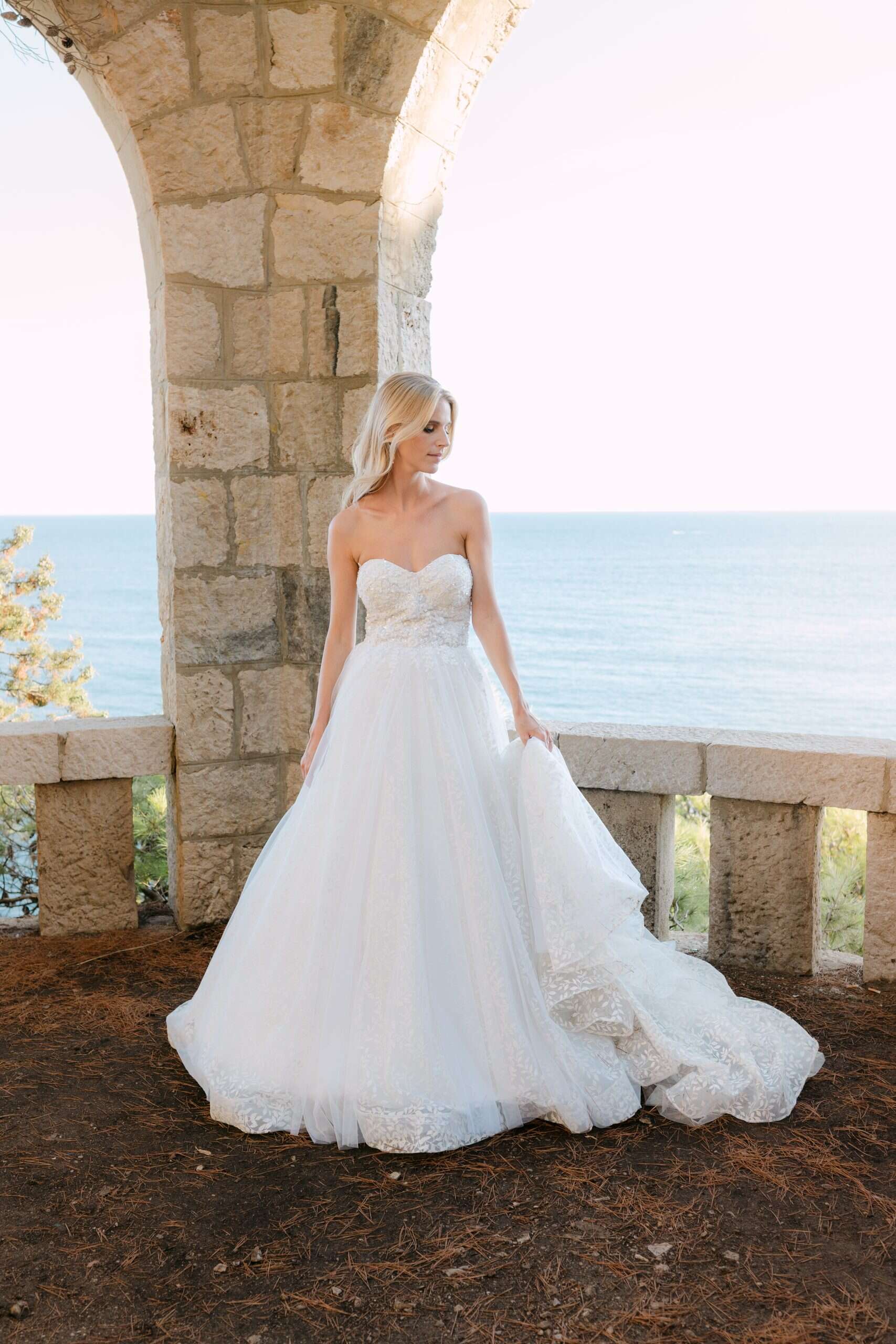 Prekrasna mladenka stoji ispred povijesnog kamenog zida u Dubrovniku, drži svoju vjenčanicu i gleda u daljinu. Iza nje se prostire predivan pogled na plavo more.