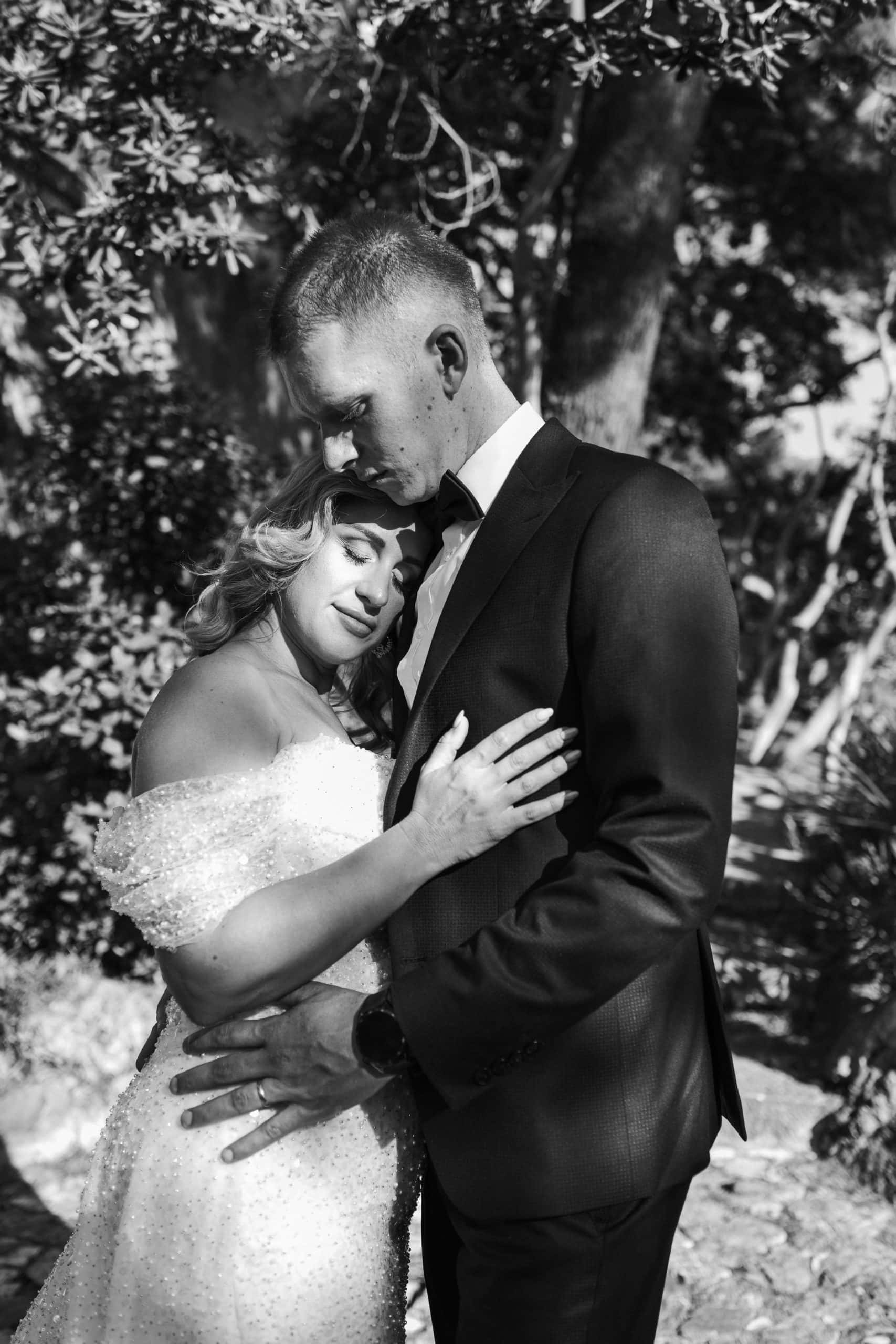 Crno-bijeli portret zagrljaja mladenaca. Mladenka nježno naslanja glavu na mladoženju u mediteranskom okruženju borova, s naglašenim filmskim zrnom.