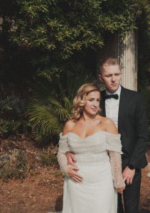 Editorial portret mladenaca obasjanih toplim svjetlom s bogatim tamnim zelenilom u pozadini. Mladoženja stoji iza mladenke i nježno je drži za struk.