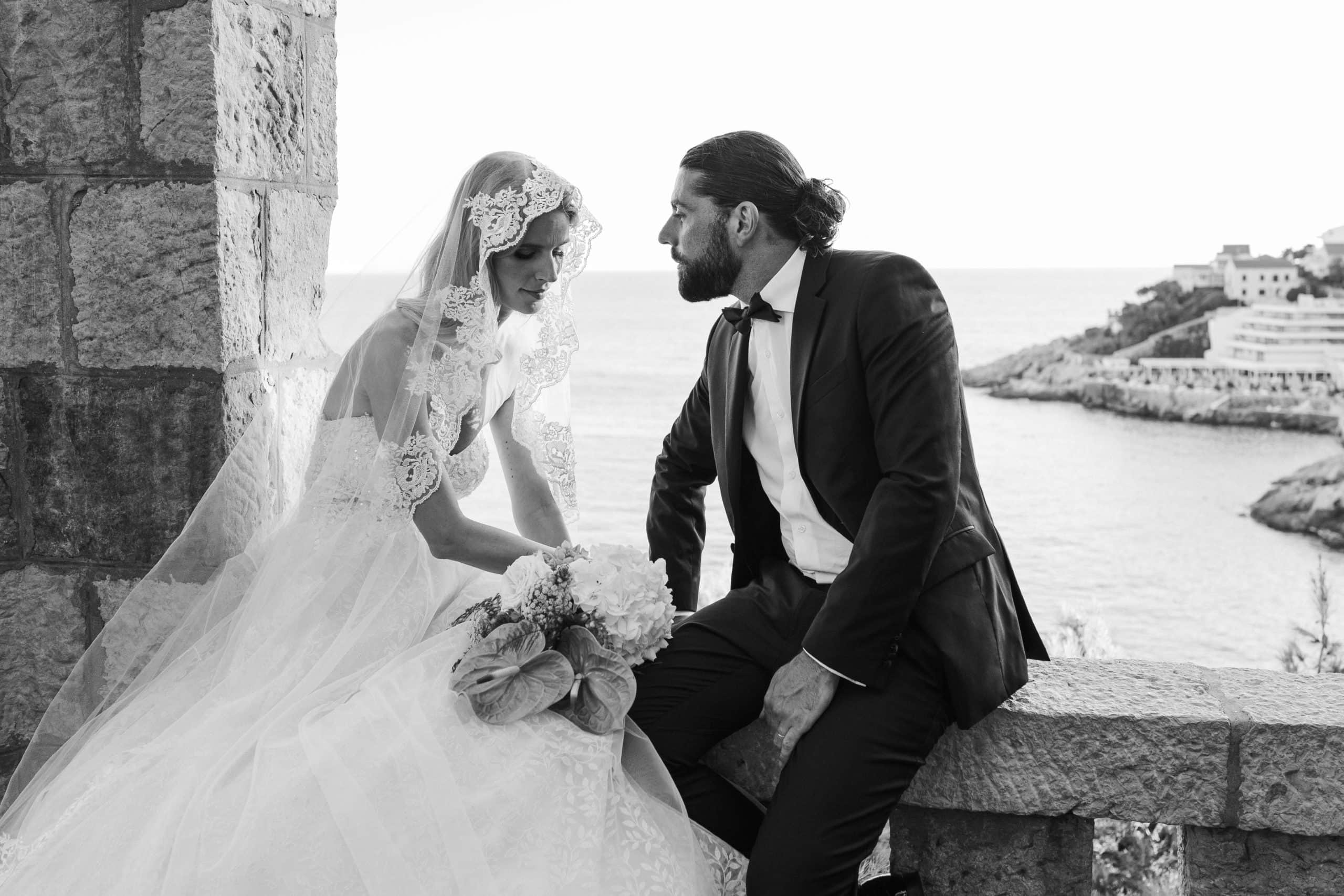 Crno-bijela fotografija mladenaca koji sjede na kamenoj ogradi iznad mora. Mladenka nosi predivan čipkasti veo i drži buket dok zaljubljeno mladoženja zaljubljeno gleda u nju.