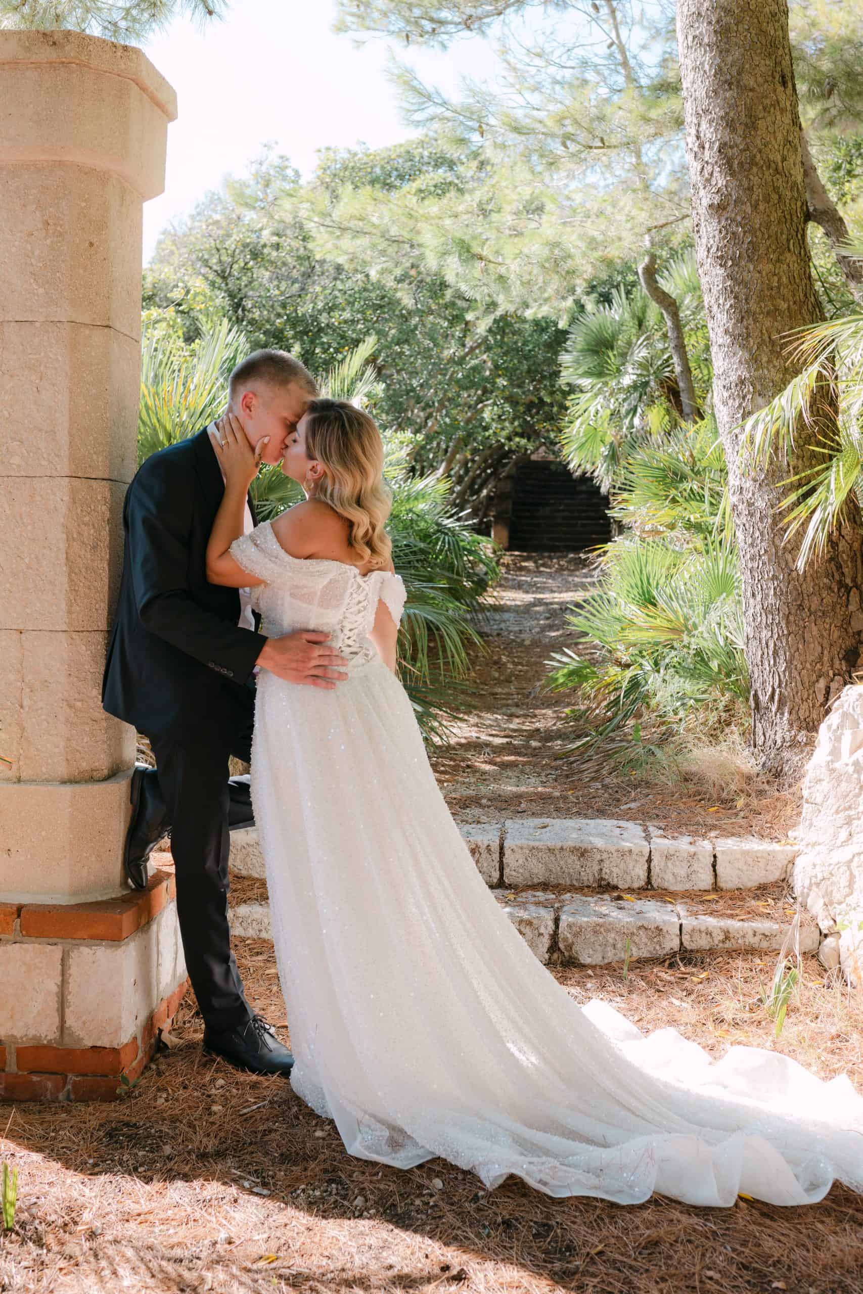 Mladenci se nježno ljube dok stoje pored velikog kamenog stupa. Oko njih je mediteransko zelenilo i kamene stepenice okružene borovima.