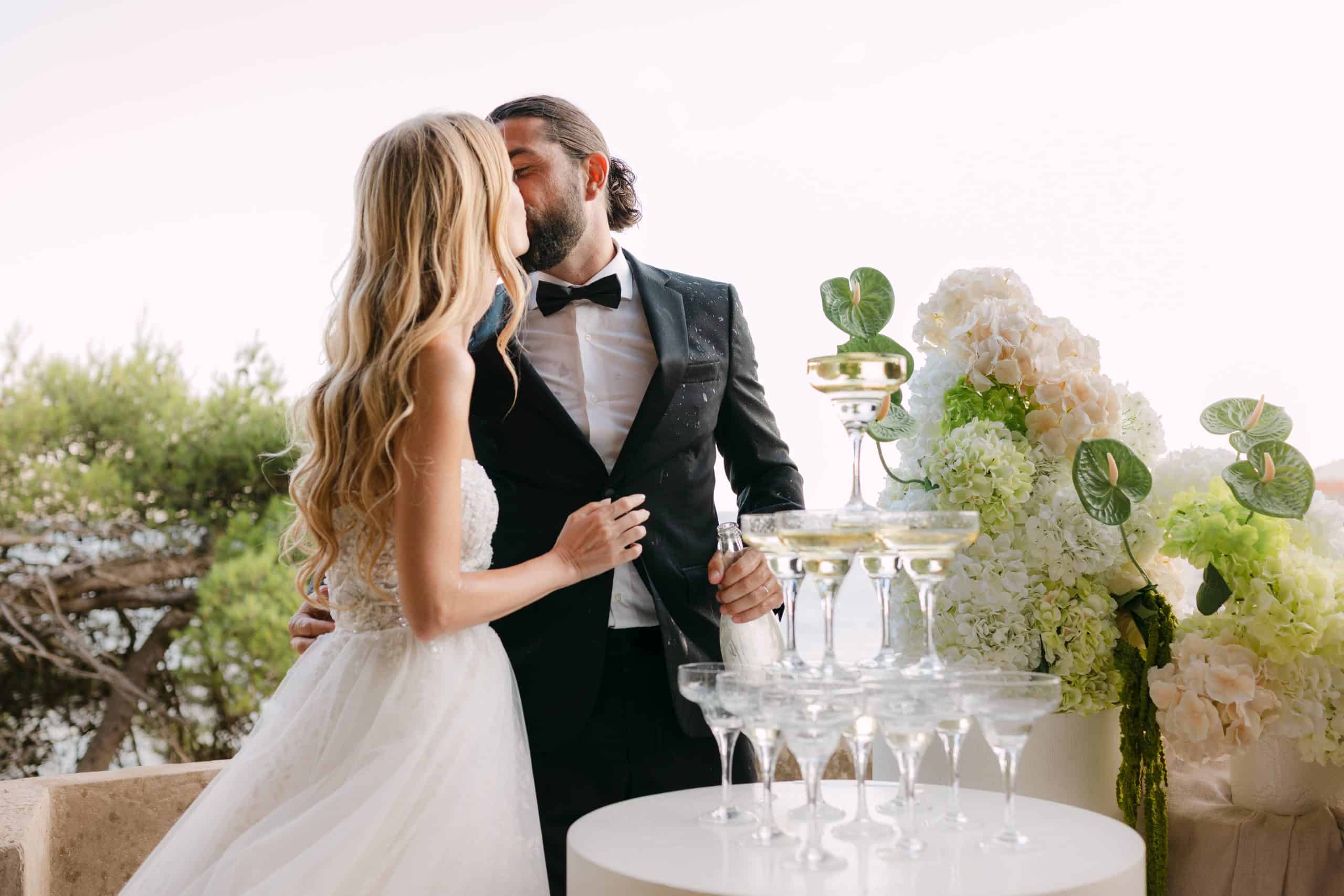 Romantični poljubac mladenaca iza kule od šampanjca. Mladoženja u ruci drži bocu, dok su okruženi predivnim cvjetnim aranžmanima na terasi u Dubrovniku.