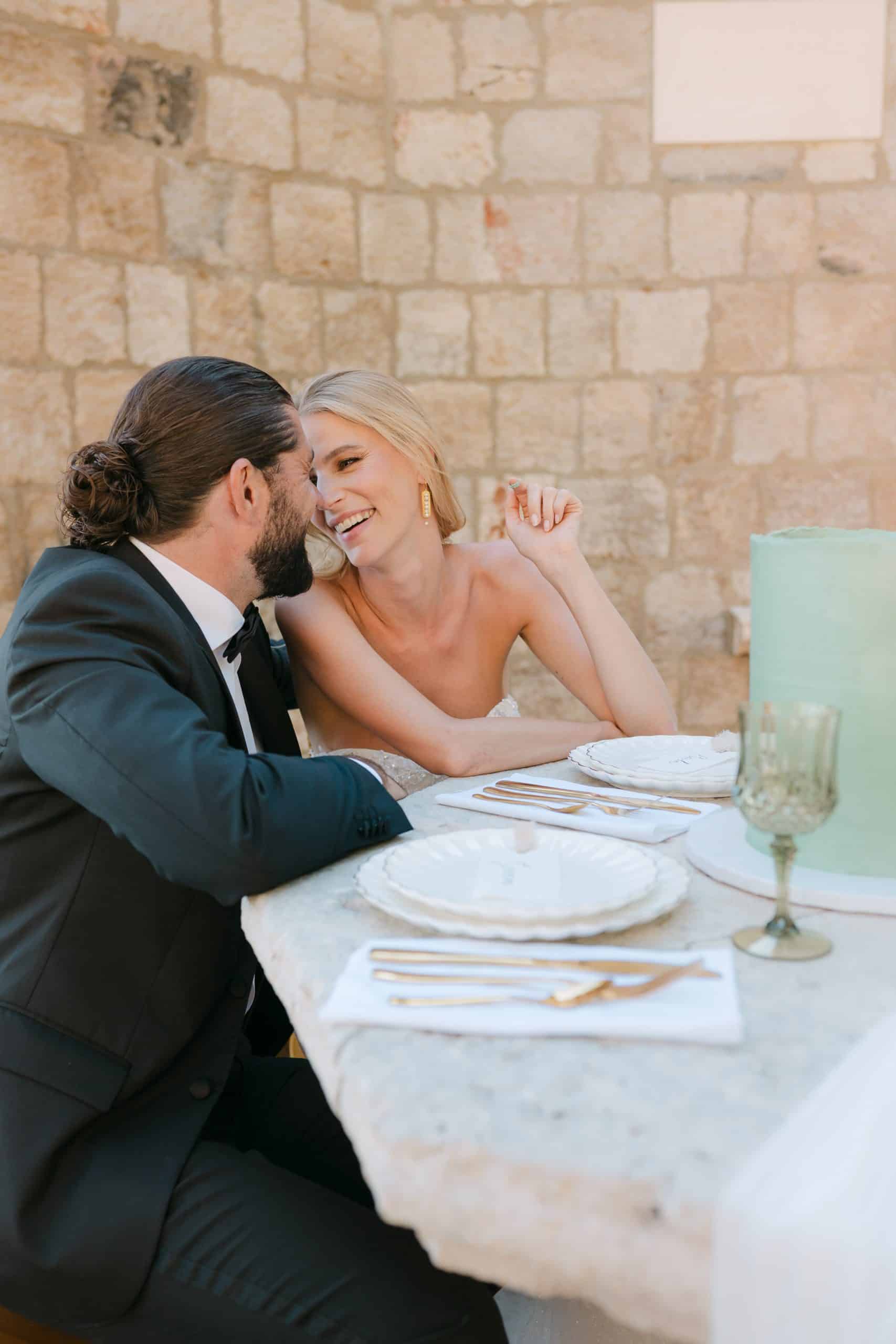 Opušteni i iskreni trenuci mladenaca koji sjede za bogato dekoriranim svadbenim stolom. Nasmiješeni par zaljubljeno se gleda tijekom proslave destination vjenčanja.