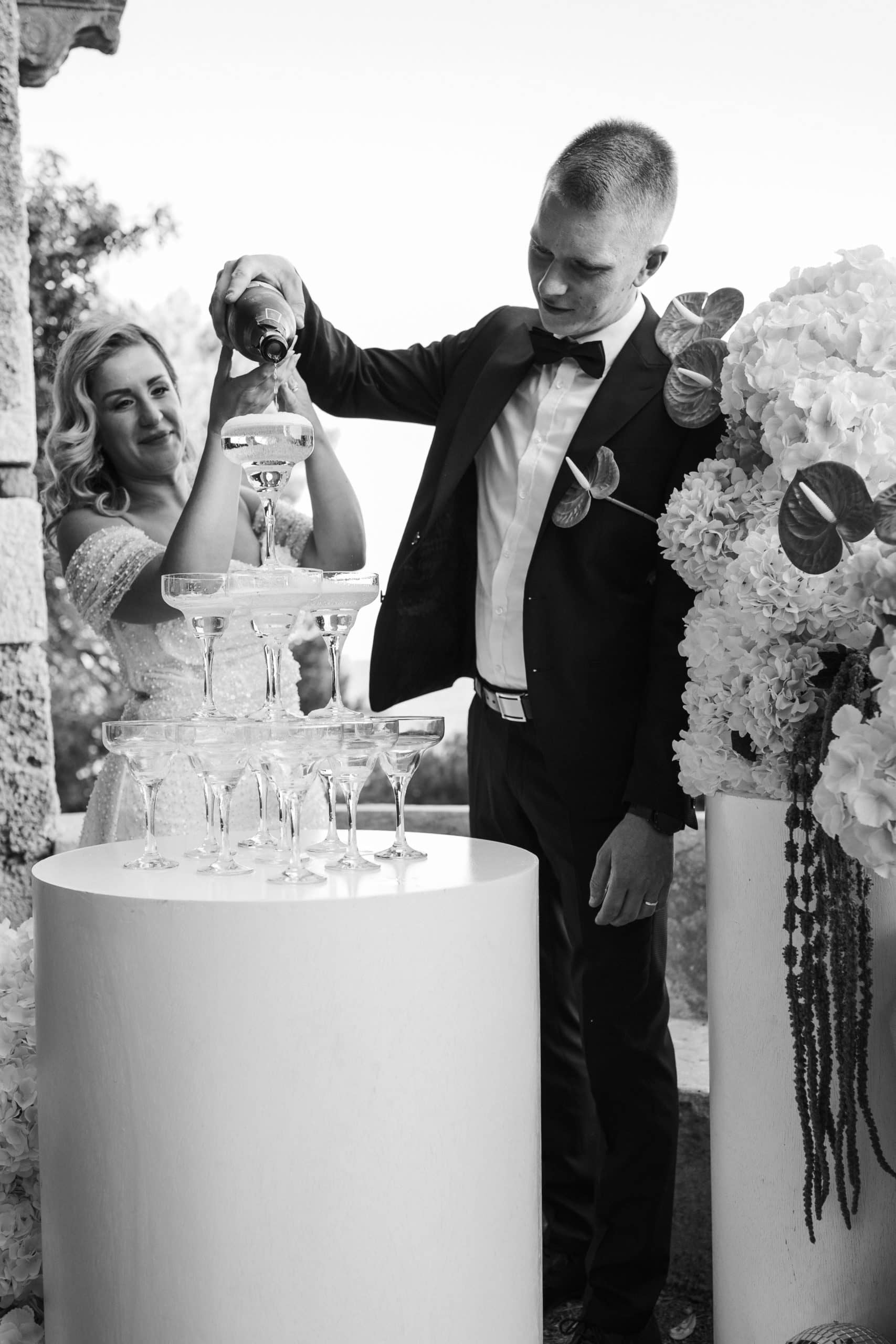 Crno-bijela fotografija mladenaca koji zajednički drže bocu i toče šampanjac na vrh staklene kule od čaša. Veseli i slavljenički trenutak na terasi u Dubrovniku.