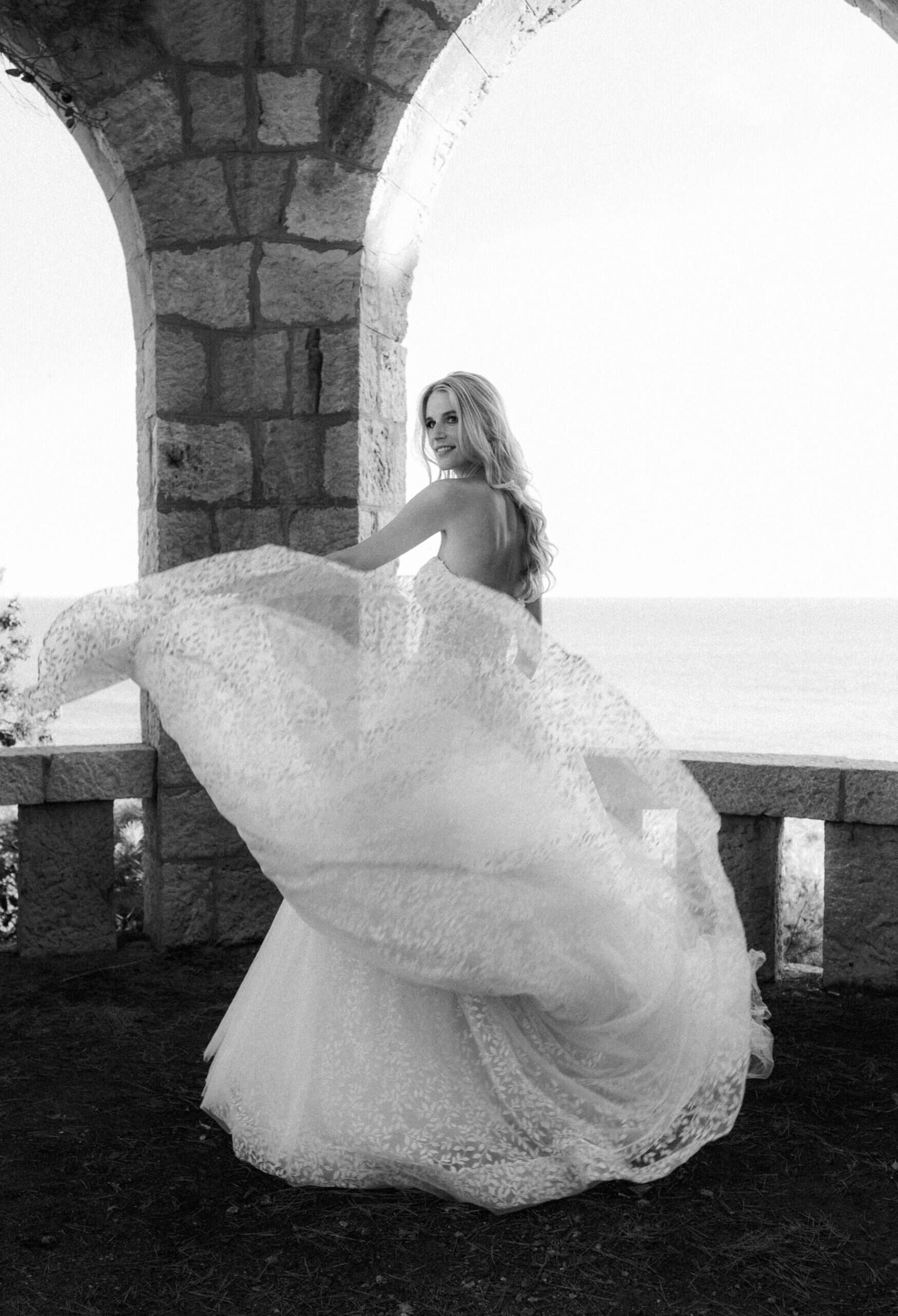 Filmska crno-bijela fotografija mladenke u pokretu. Mladenka se okreće prema kameri dok se njezin dugi čipkasti veo predivno širi i leprša ispod povijesnog luka.