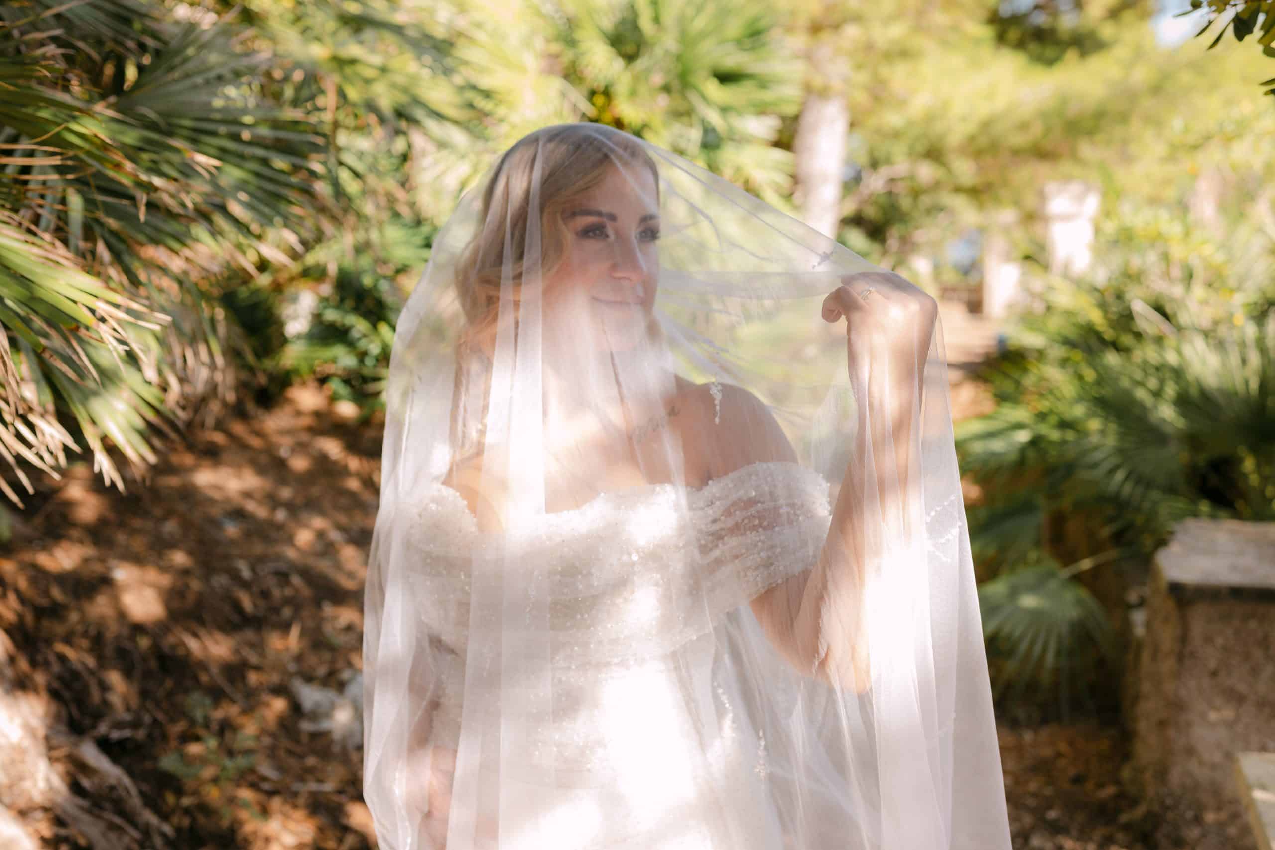 Nasmiješena mladenka u raskošnoj vjenčanici drži veo preko lica dok stoji obasjana suncem u zelenom mediteranskom vrtu Vile Čingrija.
