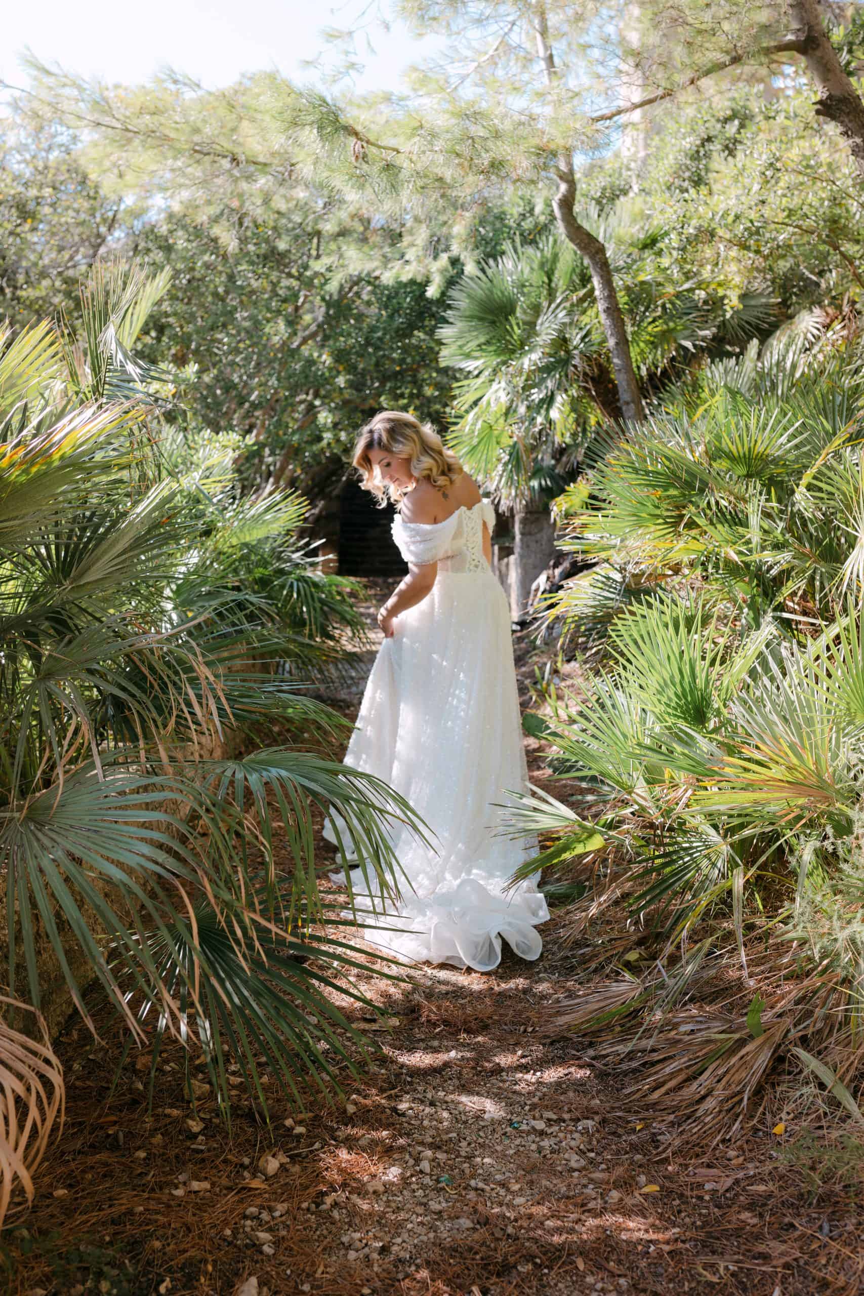 Mladenka u raskošnoj vjenčanici šeta zemljanom stazom okružena bujnim zelenilom i palmama. Sunčan ljetni dan na destination vjenčanju u Dubrovniku.