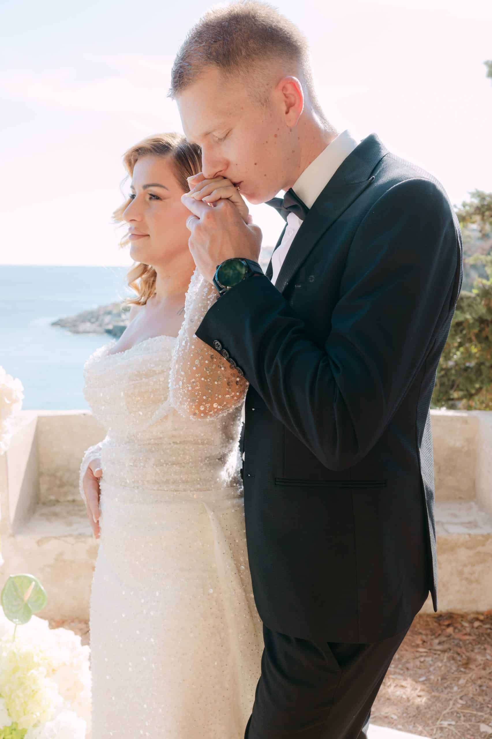 Profilni portret mladenaca na sunčanom balkonu s pogledom na more. Mladoženja nježno ljubi ruku mladenki tijekom destination vjenčanja u Dubrovniku.