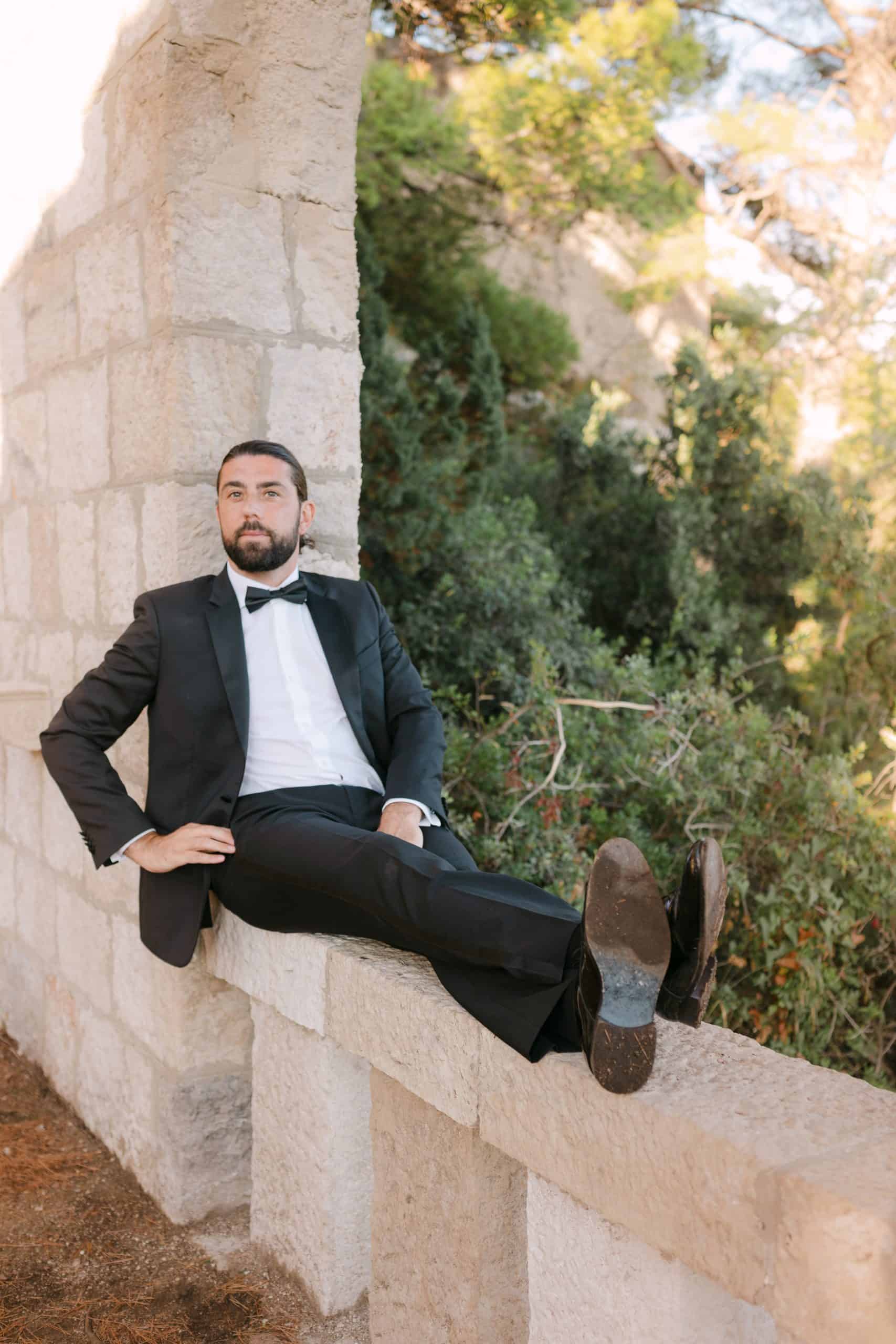 Ležeran i opušten portret mladoženje u crnom odijelu s leptir mašnom. Mladoženja sjedi na kamenoj ogradi ispruženih nogu, okružen mediteranskim zelenilom.