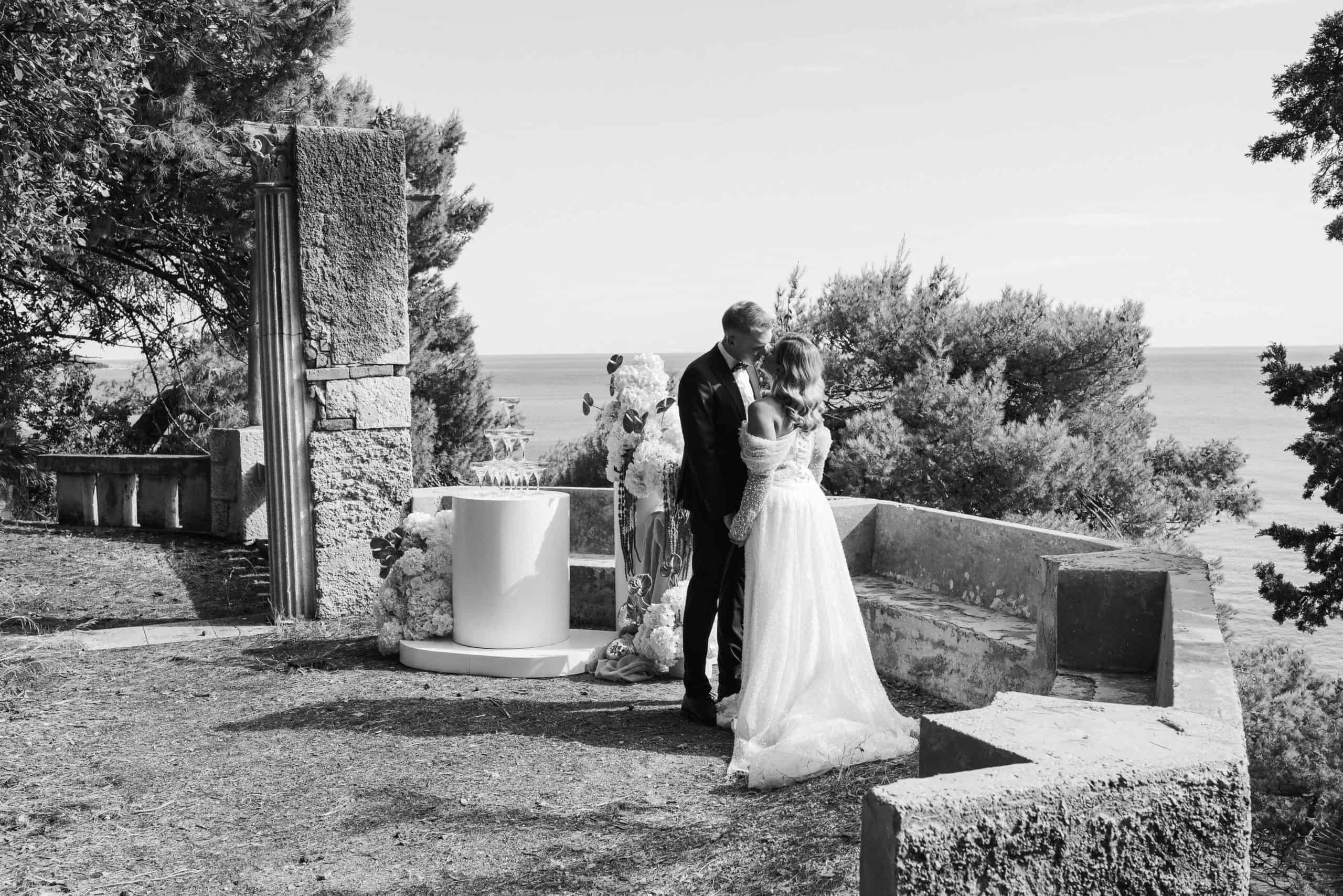 Crno-bijela fotografija romantičnog poljupca mladenaca ispred svadbene dekoracije i kule od šampanjca na kamenoj terasi s pogledom na more.