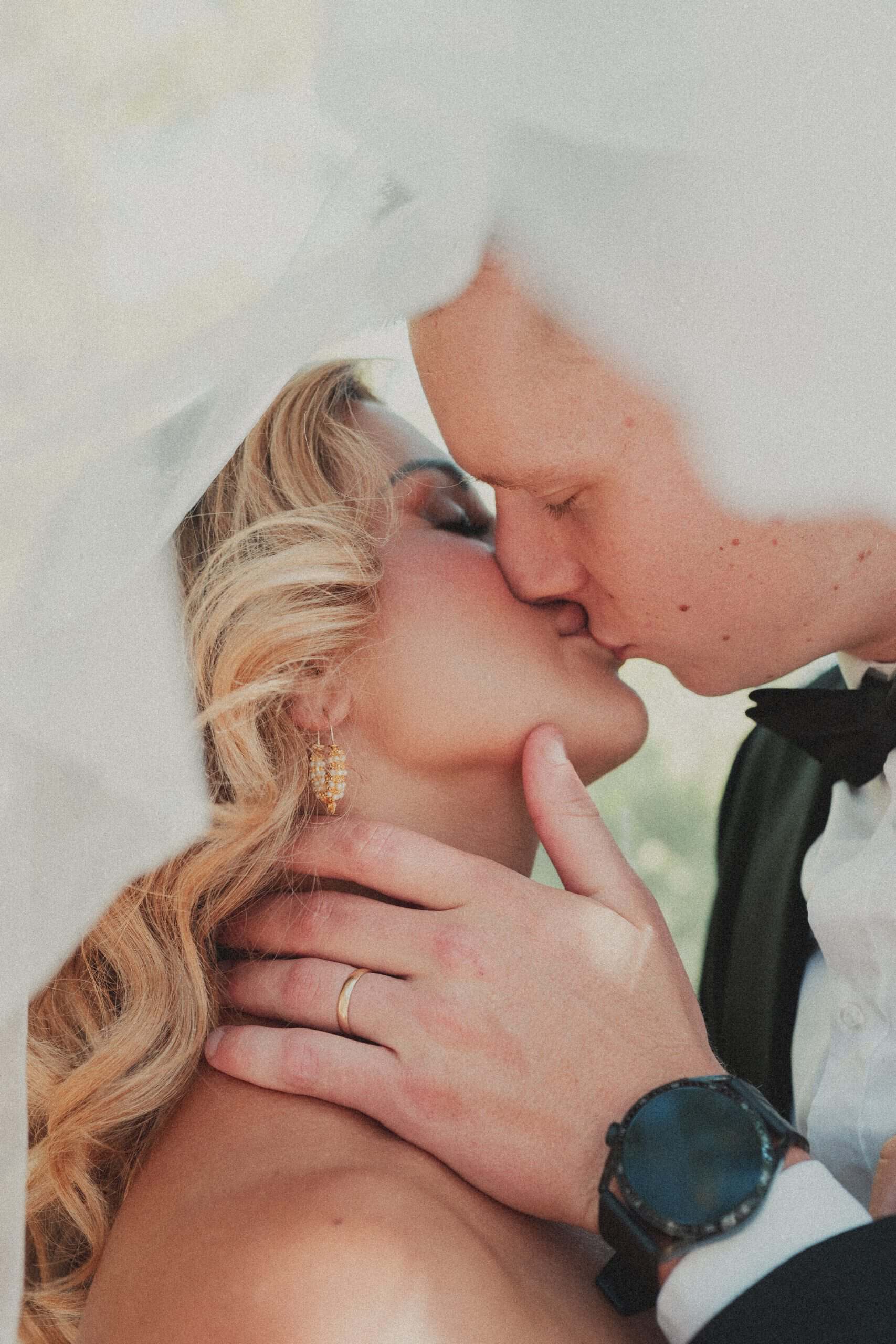 Krupni kadar romantičnog poljupca mladenaca ispod mladenkinog bijelog vela. Topli tonovi i intimna atmosfera svadbenog sessiona.