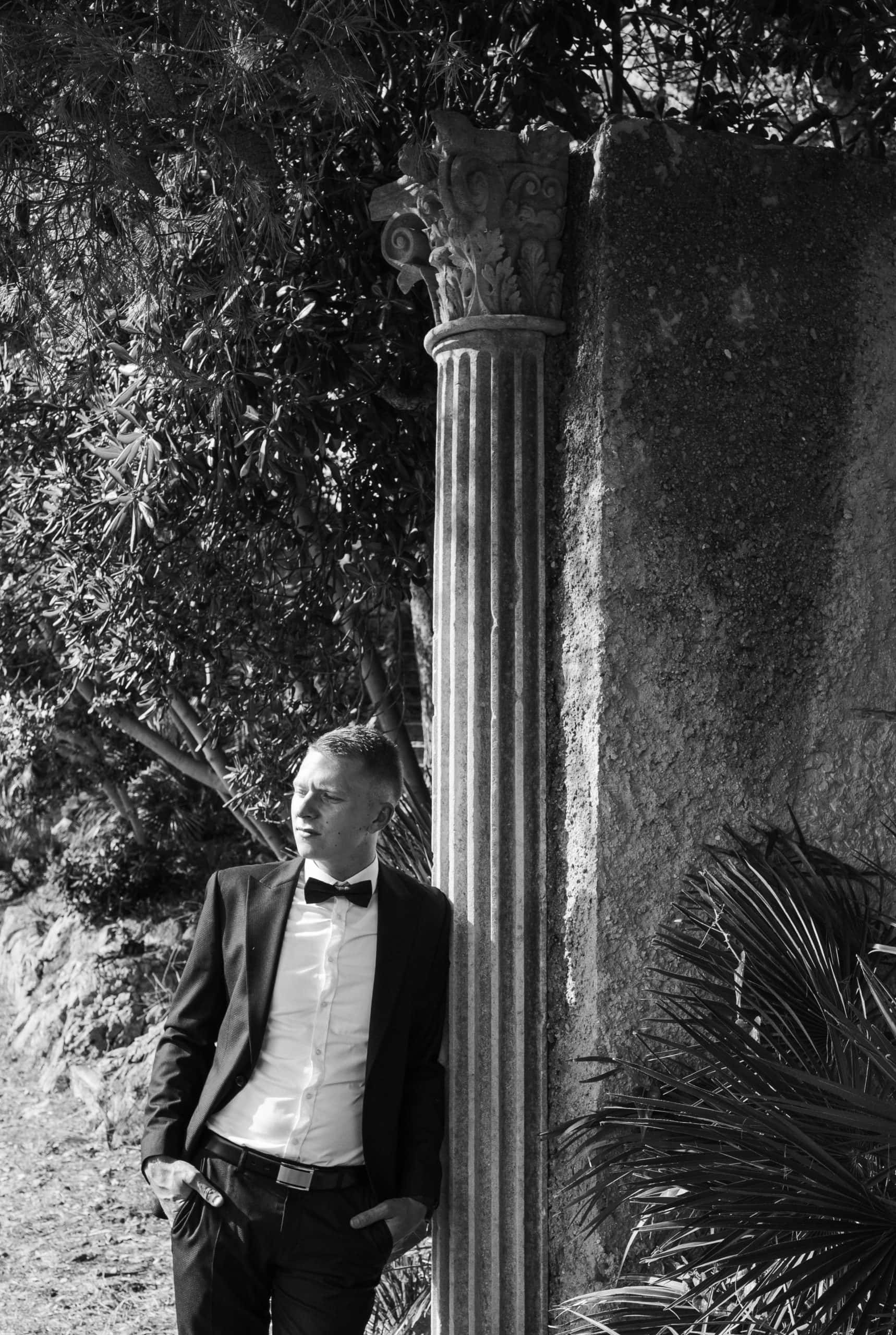 Crno-bijeli portret mladoženje koji zamišljeno gleda u daljinu, ležerno naslonjen na povijesni kameni stup u hladovini drveća.