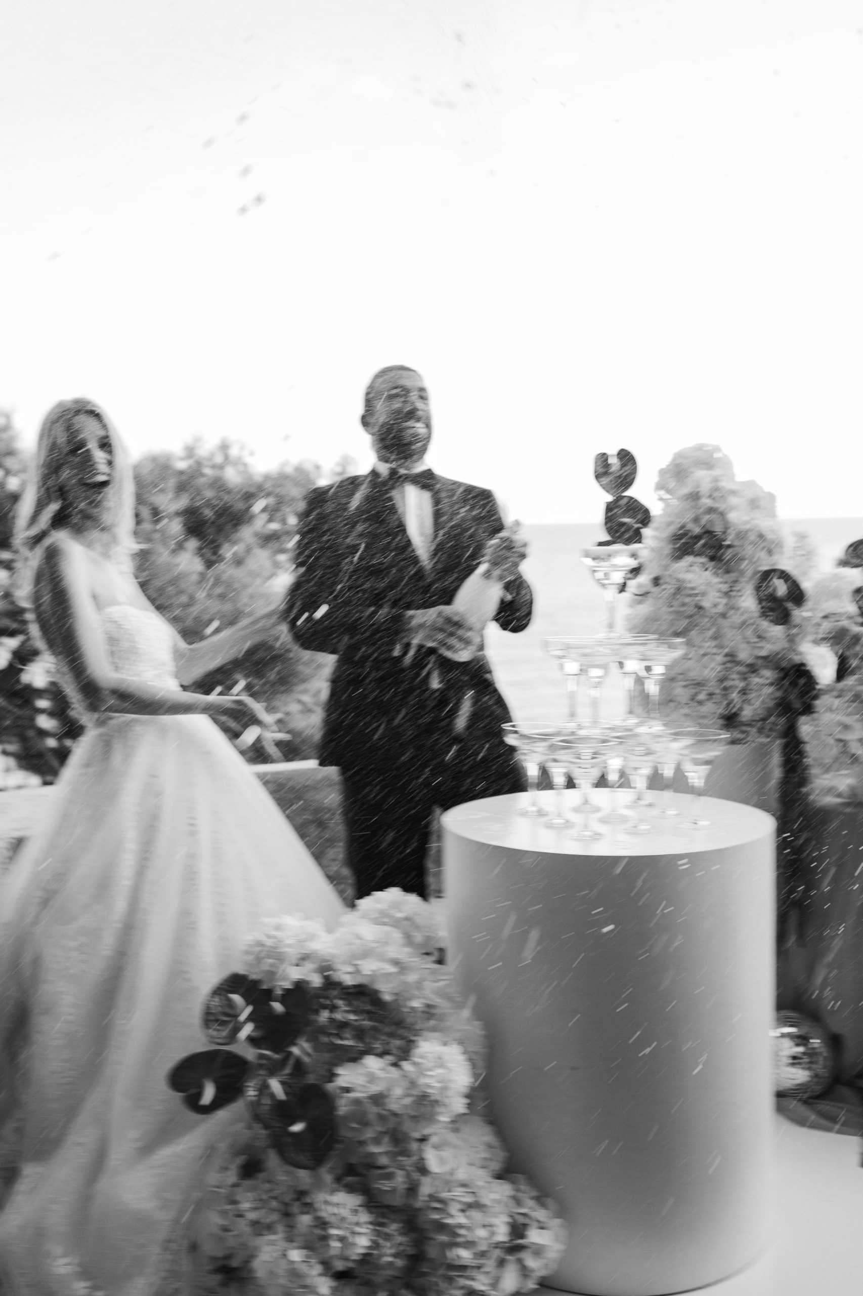 Dokumentarna crno-bijela fotografija slavlja. Mladoženja veselo prska šampanjac u zrak dok se mladenka smije pored kule od čaša tijekom destination vjenčanja.