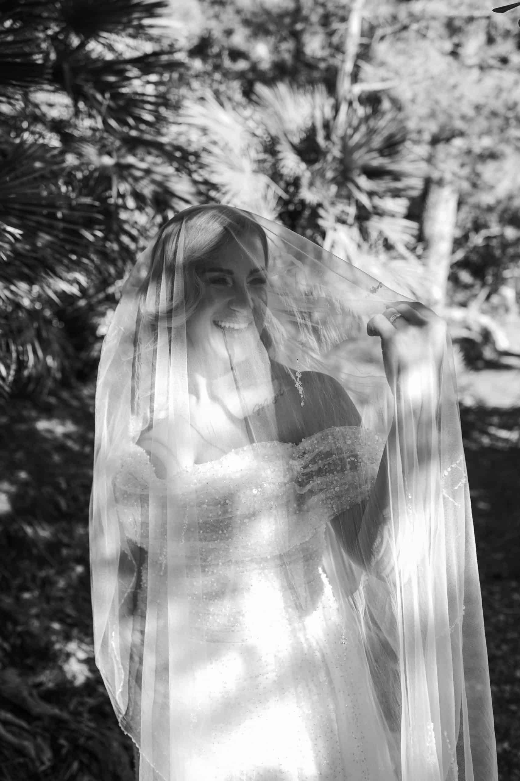Crno-bijeli portret široko nasmiješene mladenke ispod vela. Prekrasna igra svjetla i sjene u prirodi stvara filmski ugođaj.