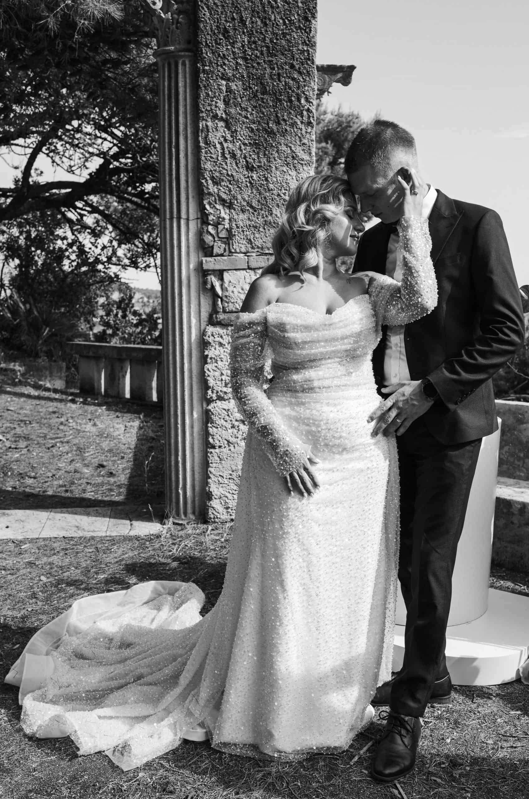 Intimni crno-bijeli kadar mladenaca. Mladoženja nježno dodiruje lice mladenke dok poziraju okruženi prirodom i povijesnom kamenom arhitekturom.