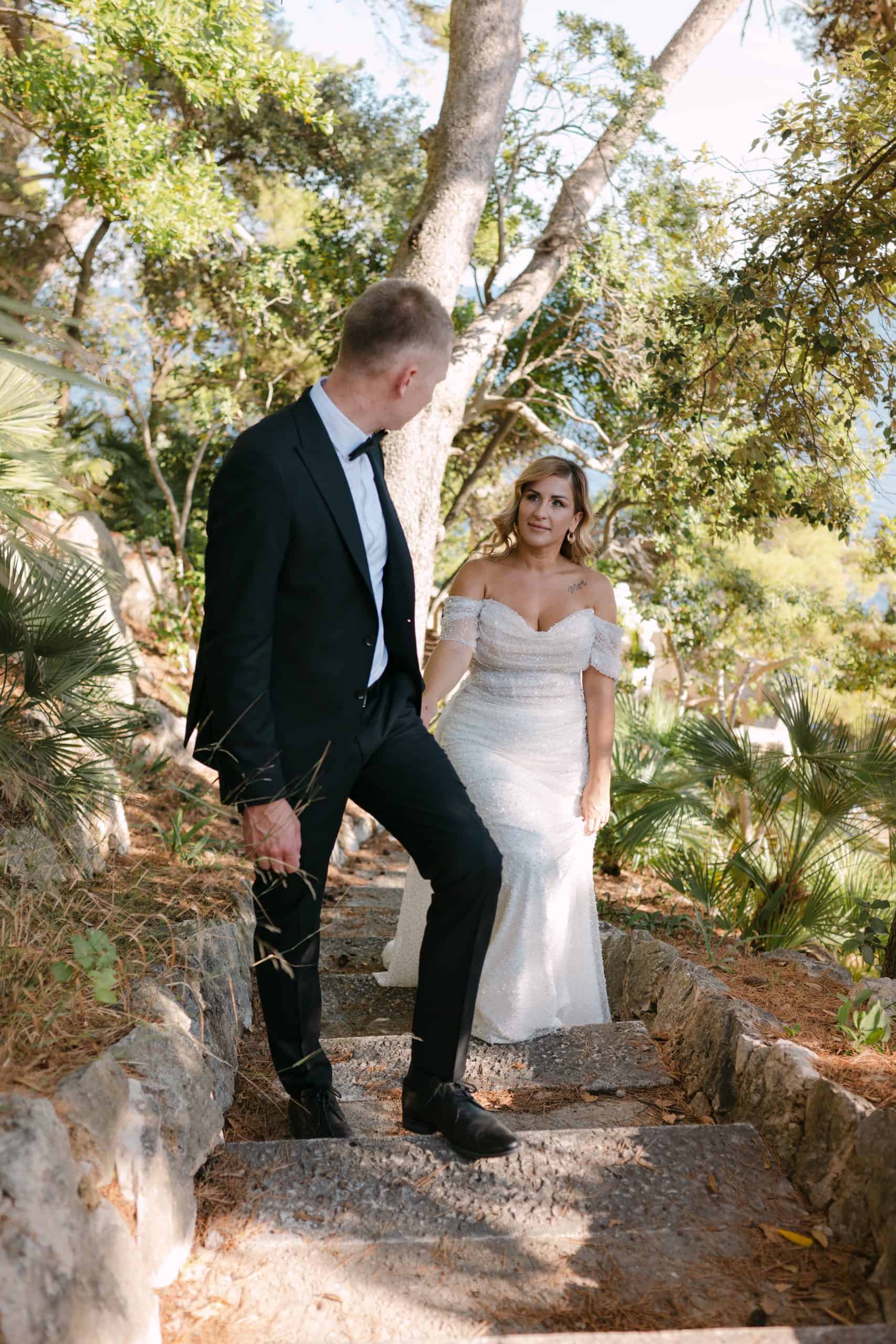 Spontani trenutak mladenaca u šetnji kamenim stepenicama. Mladoženja stoji stepenicu više i gleda u mladenku u raskošnoj vjenčanici. Mediteranski vrt u Dubrovniku.