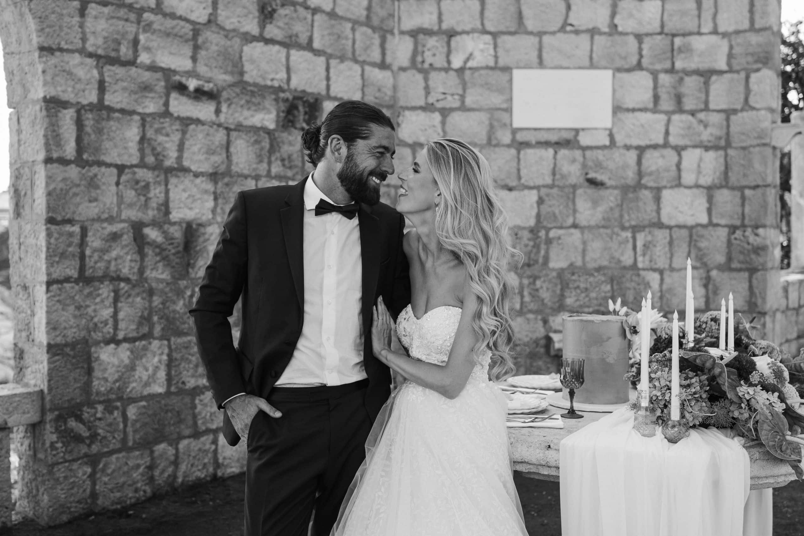 Opušteni i nasmiješeni mladenci prepuni emocija gledaju se pored bogato dekoriranog svadbenog stola s tortom, svijećama i cvijećem. Crno-bijela svadbena fotografija ispred povijesnog kamenog zida u Dubrovniku.