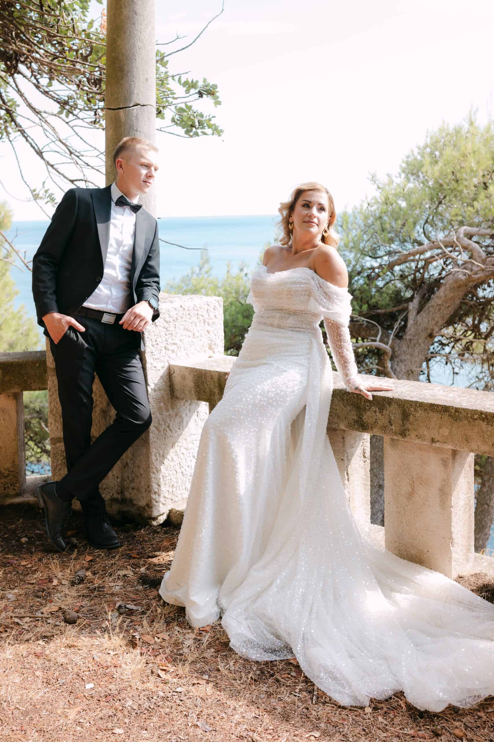 Mladenci poziraju na osunčanoj kamenoj terasi s predivnim pogledom na more. Mladoženja je ležerno naslonjen na stup, dok mladenka u raskošnoj svjetlucavoj vjenčanici sjedi na kamenoj ogradi tijekom destination vjenčanja u Dubrovniku.