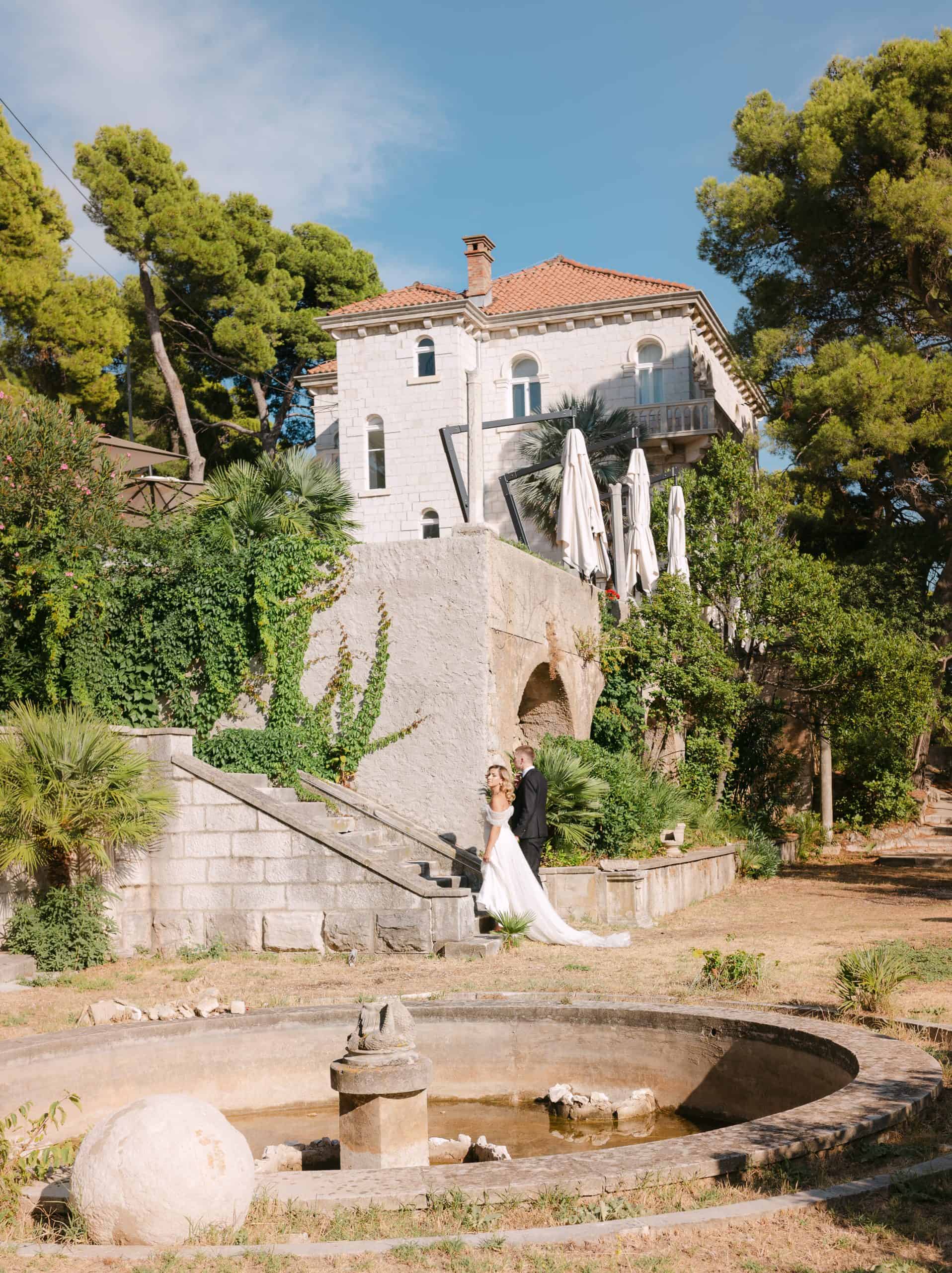 Široki kadar mladenaca koji stoje pored stare kamene fontane s pogledom na impresivnu arhitekturu Vile Čingrija. Sunčan dan i predivan mediteranski vrt u Dubrovniku.