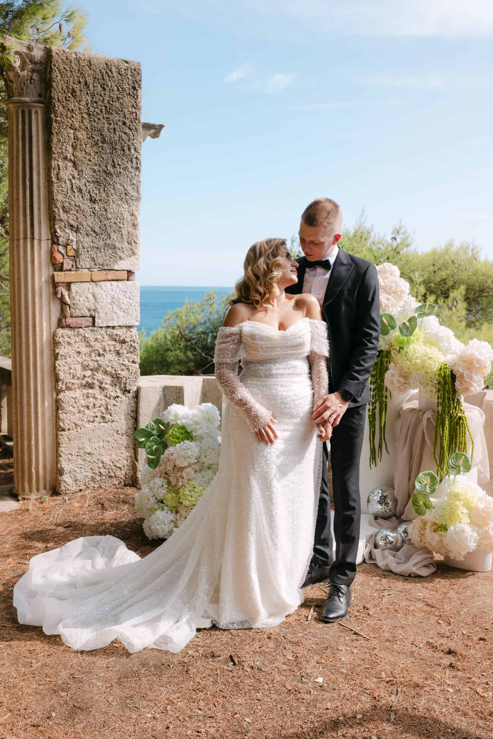 Mladenci poziraju ispred kamenog zida s pogledom na more. Mladenka nosi vjenčanicu spuštenih ramena i ima cvjetni aranžman u pozadini. Wedding session u Vili Čingrija, Dubrovnik.