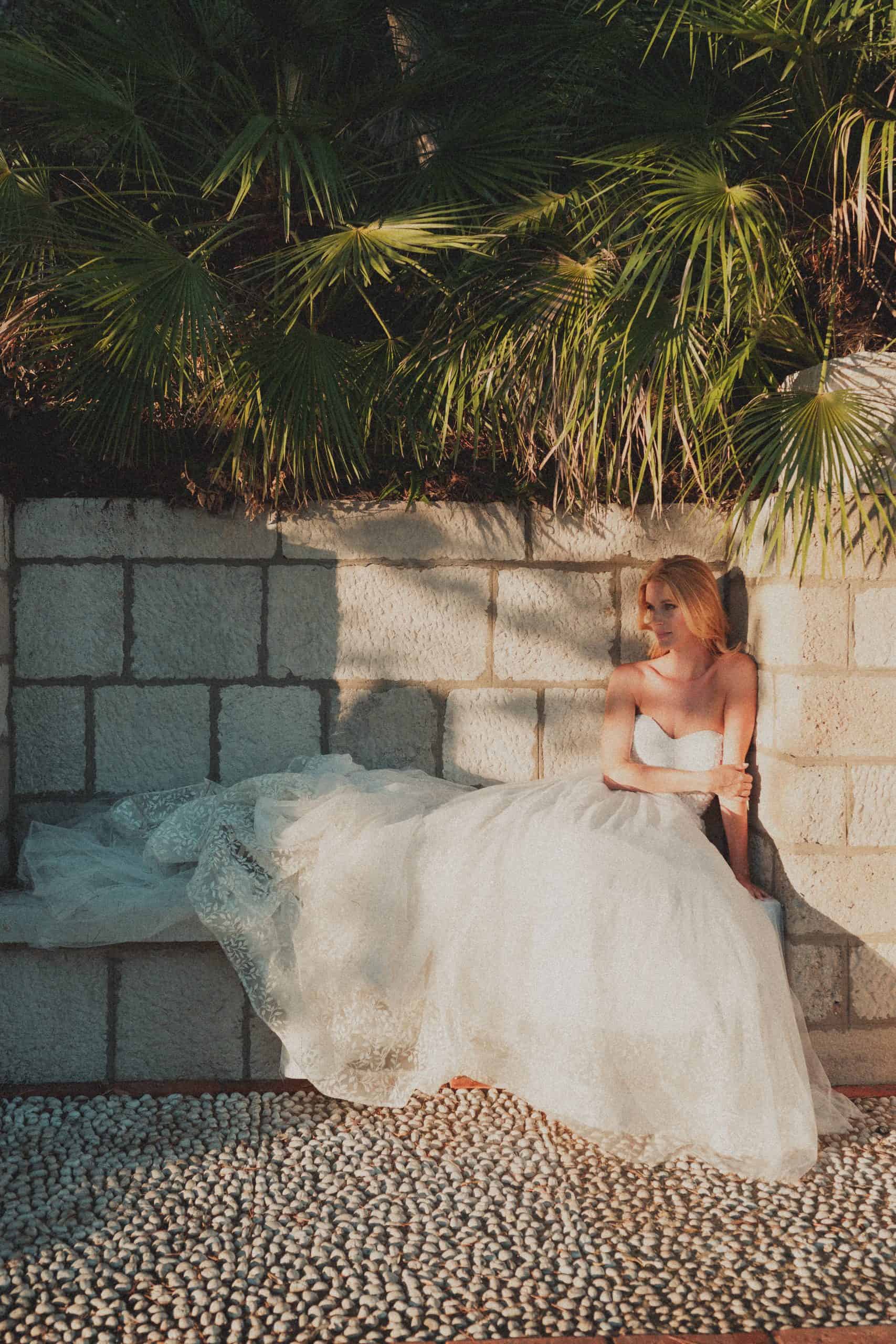 Prekrasna mladenka sjedi uz povijesni kameni zid okružena mediteranskim palmama, obasjana toplim poslijepodnevnim suncem tijekom wedding sessiona u Dubrovniku.