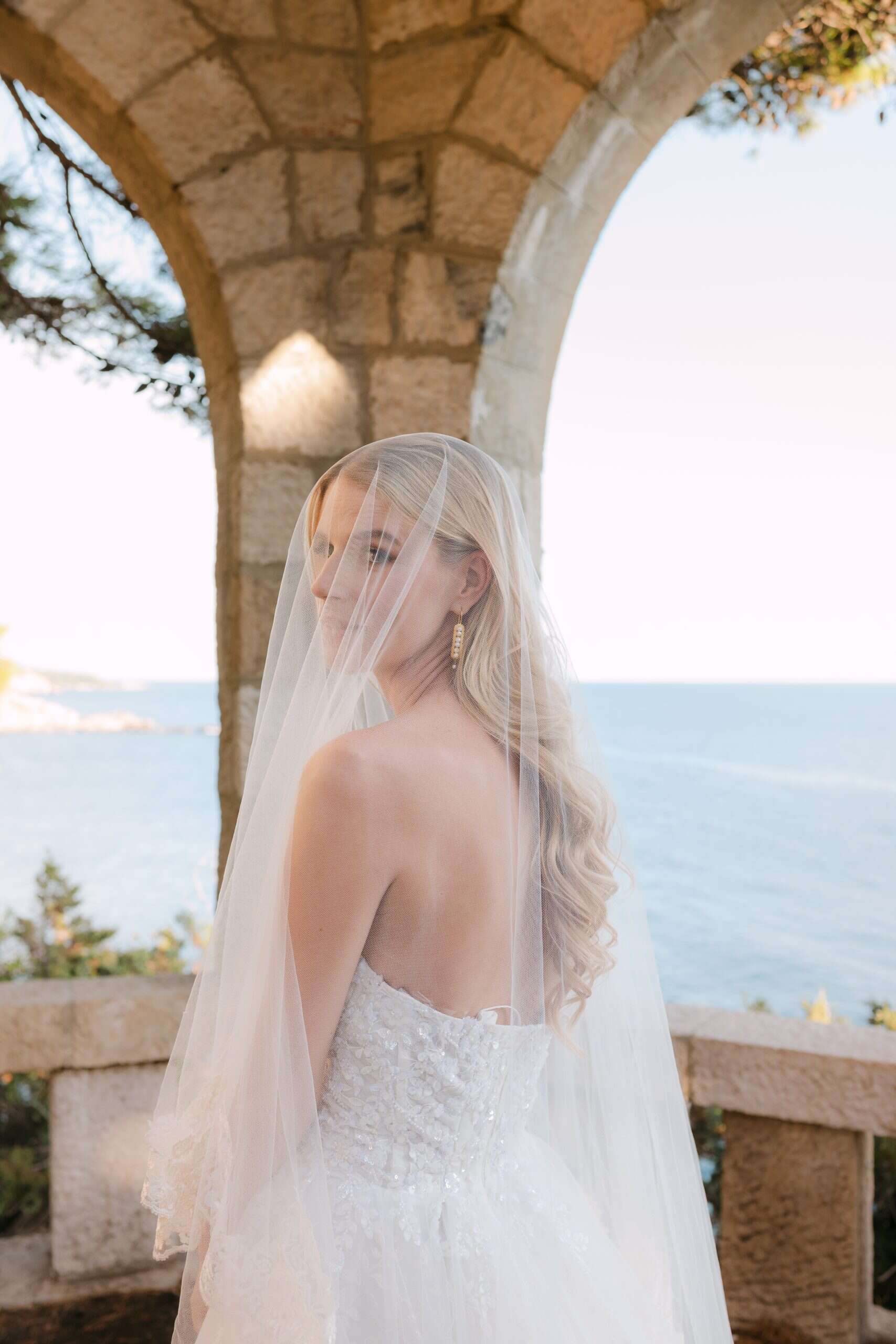 Elegantna mladenka s dugim prozirnim velom i zlatnim visećim naušnicama gleda preko ramena ispod osunčanog kamenog luka, s predivnim plavim morem u pozadini.