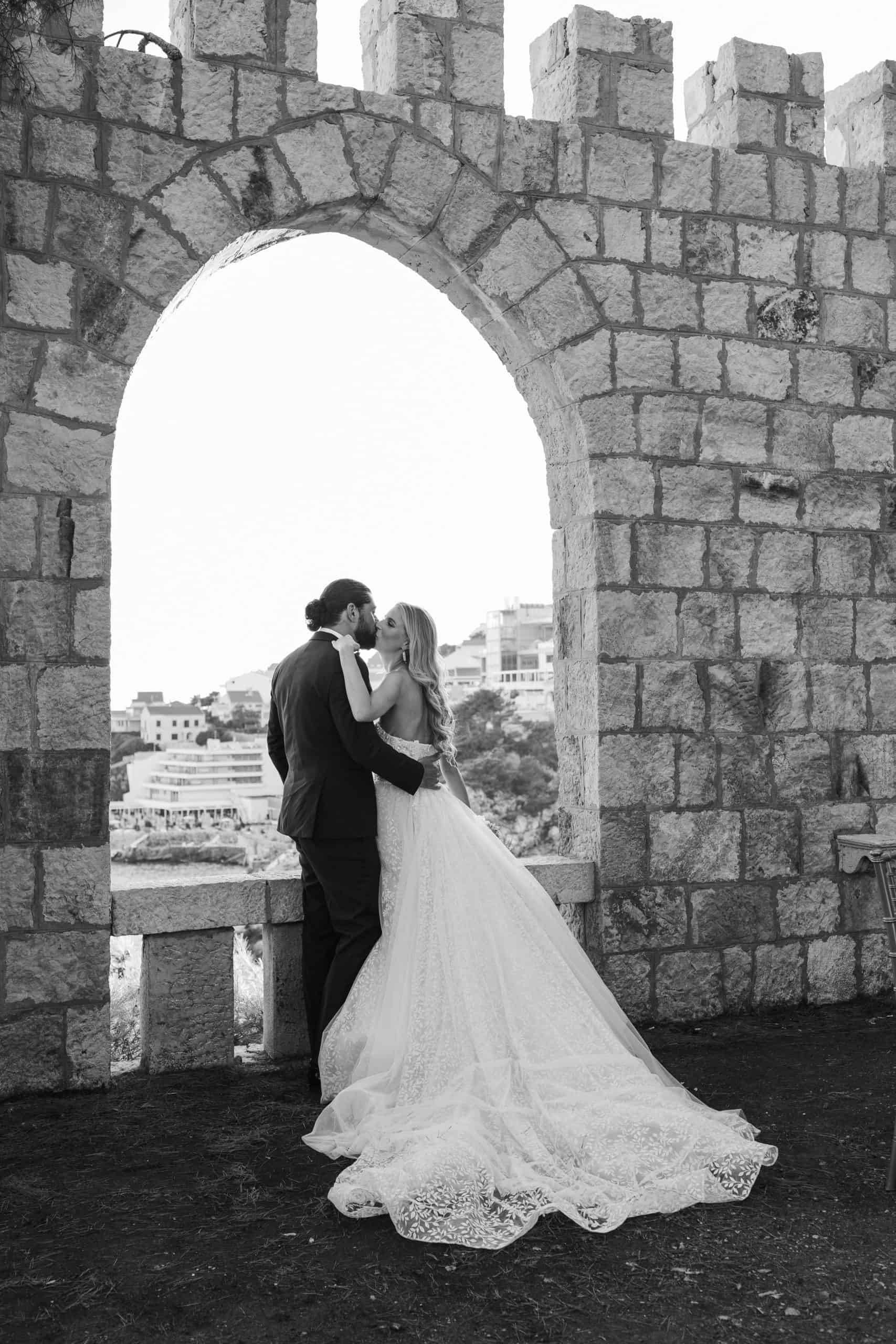 Široki crno-bijeli kadar mladenaca koji se romantično ljube ispod velikog starog kamenog luka. Bezvremenska destination wedding fotografija iz Dubrovnika.