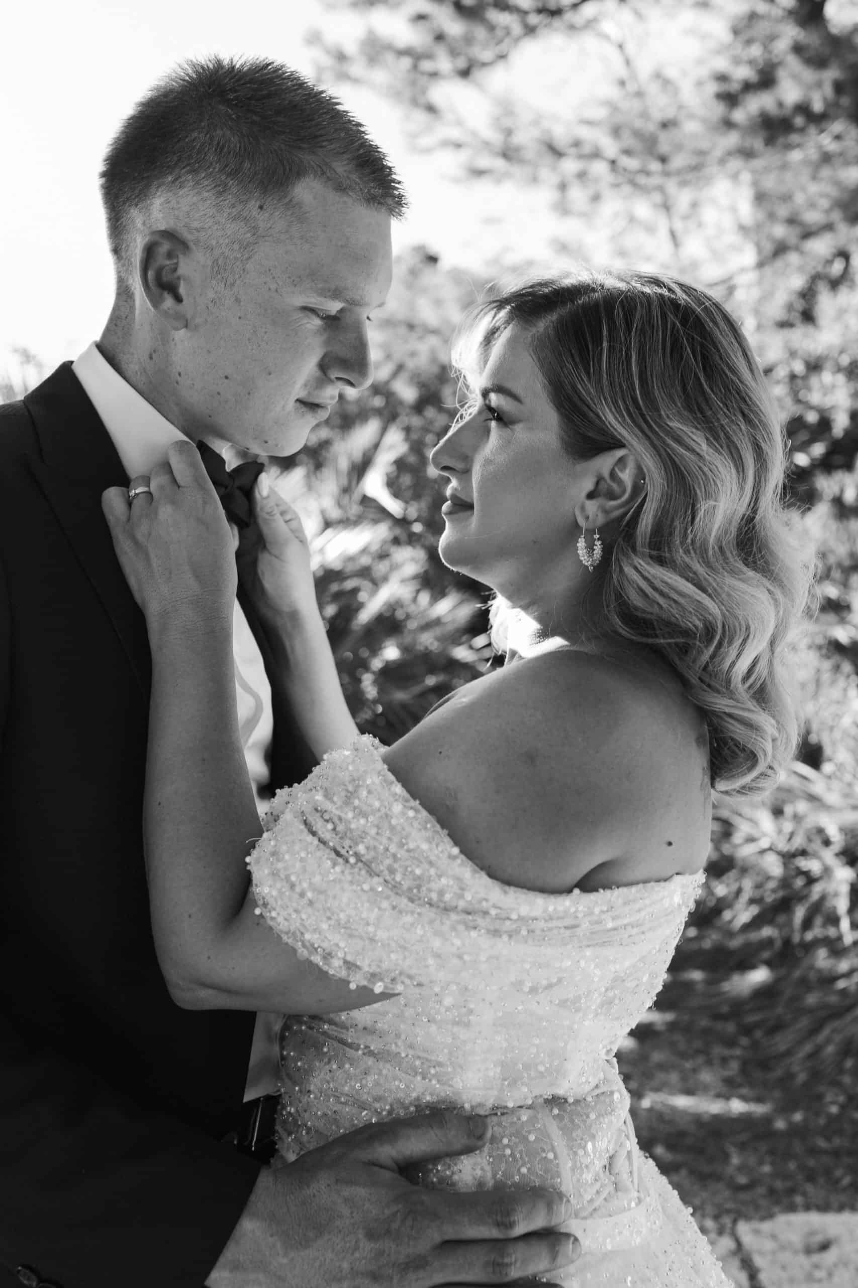 Intimni crno-bijeli portret mladenaca izbliza. Mladenka obasjana suncem gleda u mladoženju i namješta mu leptir mašnu na odijelu.