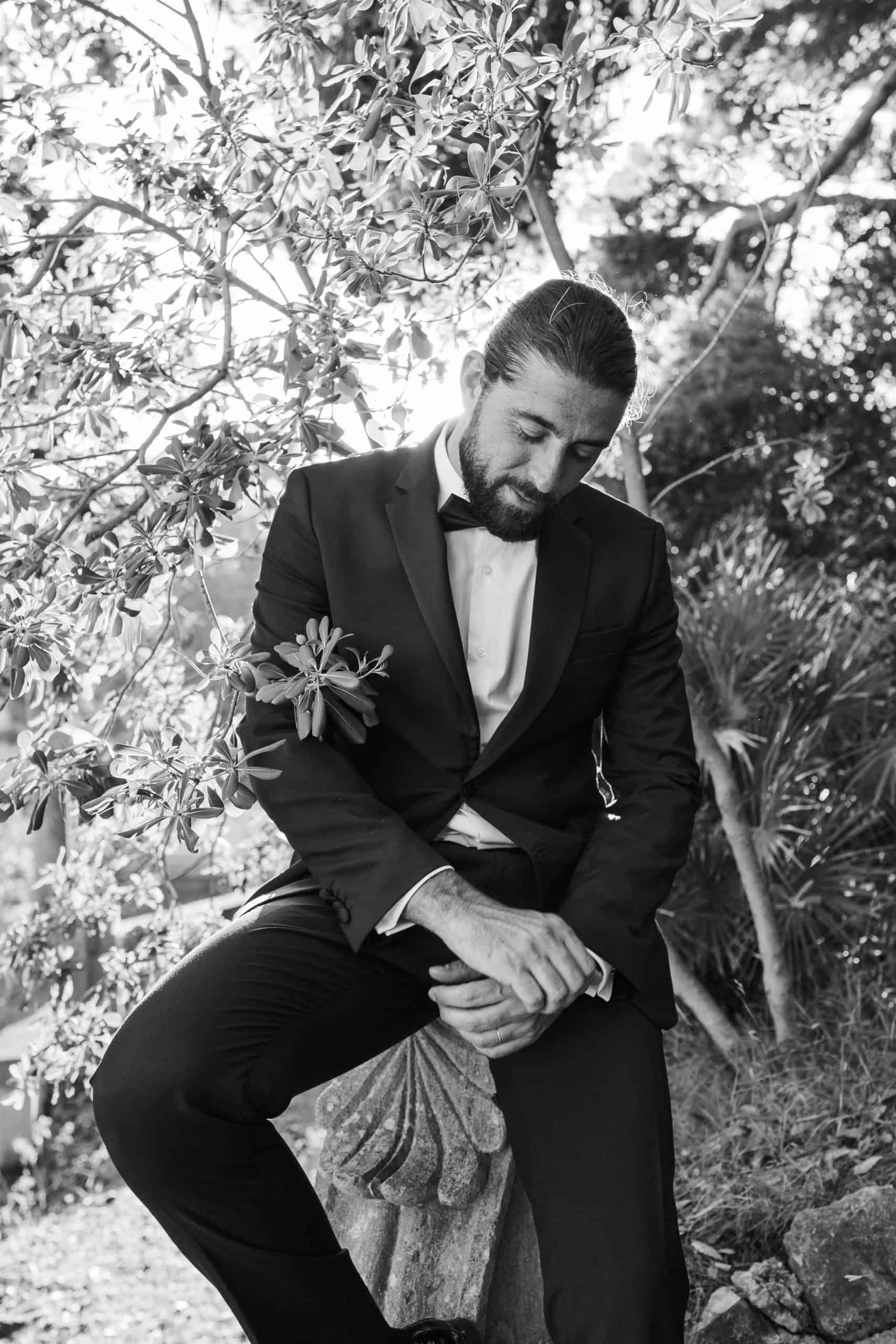 Filmski crno-bijeli portret mladoženje u elegantnom crnom odijelu s leptir mašnom. Mladoženja zamišljeno pozira okružen bogatim mediteranskim zelenilom tijekom wedding sessiona u Dubrovniku.