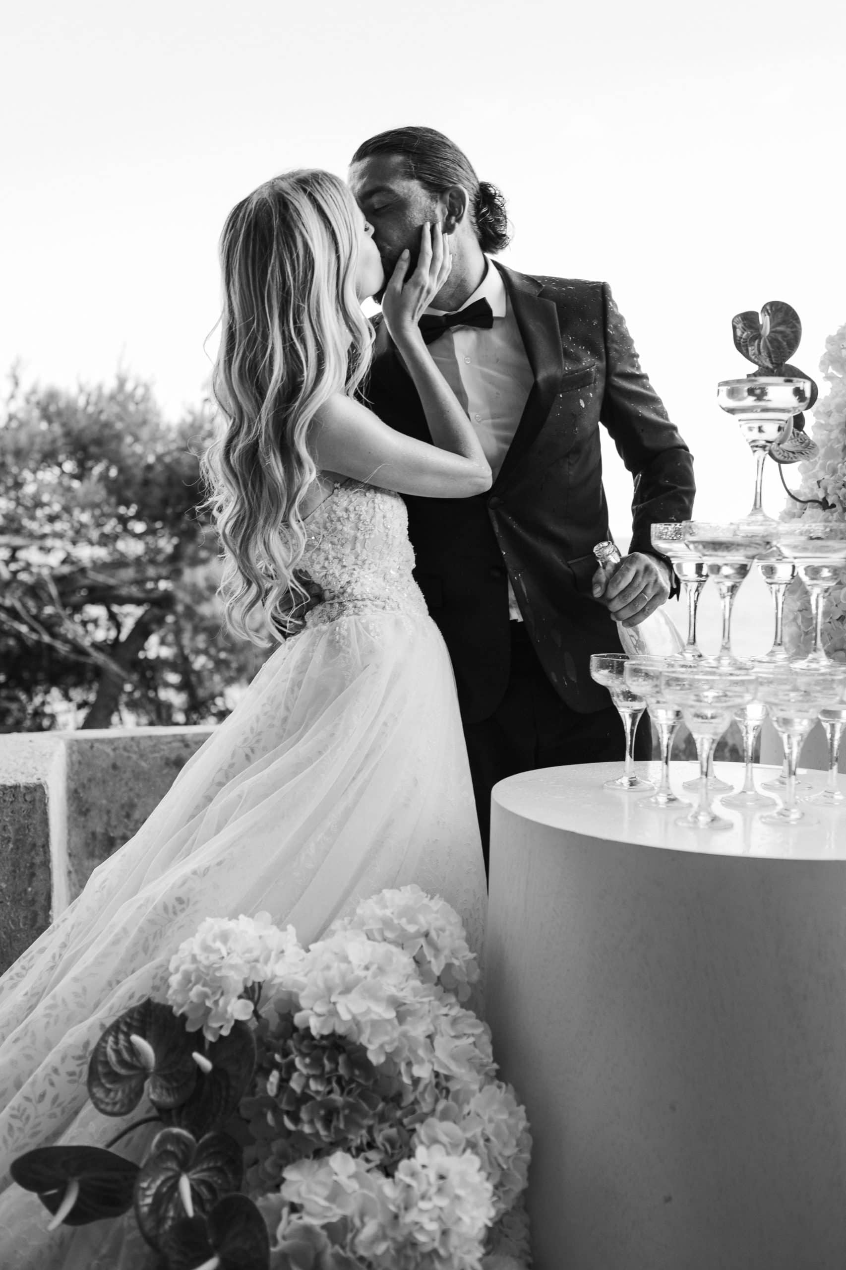 Elegantna crno-bijela svadbena fotografija mladenaca koji se ljube pored raskošne kule od čaša za šampanjac. Filmska i romantična atmosfera.