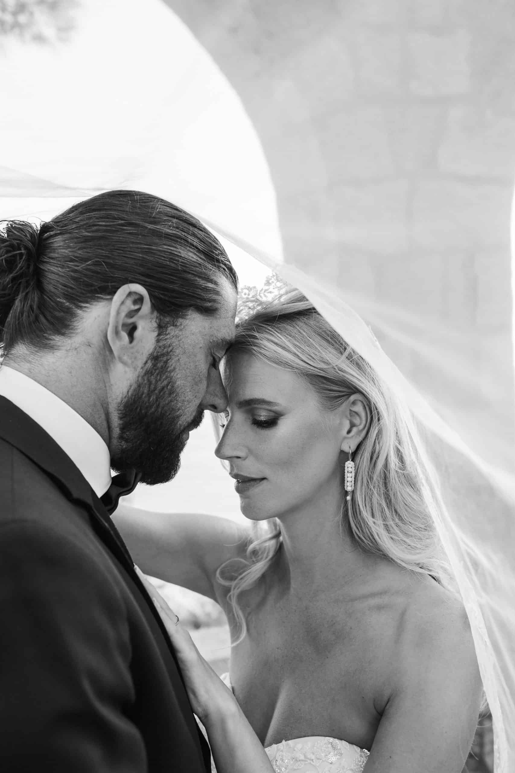 Intimni crno-bijeli krupni kadar mladenaca zatvorenih očiju. Nježno se dodiruju čelima skriveni ispod mladenkinog prozirnog vela.