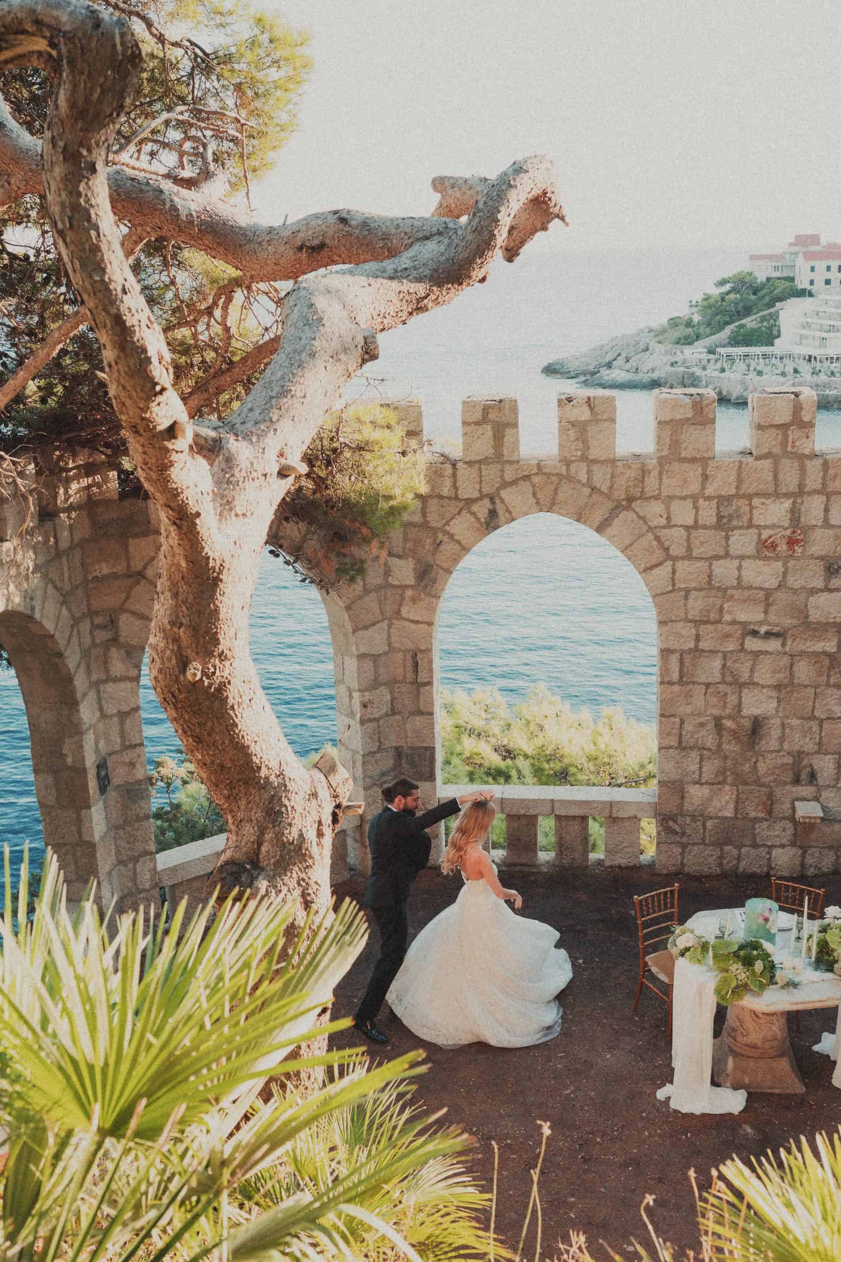 Široki kadar mladenaca koji plešu ispod povijesnog kamenog luka i velikog stabla bora, s predivnim pogledom na more. Romantična scena na destination vjenčanju u Dubrovniku.