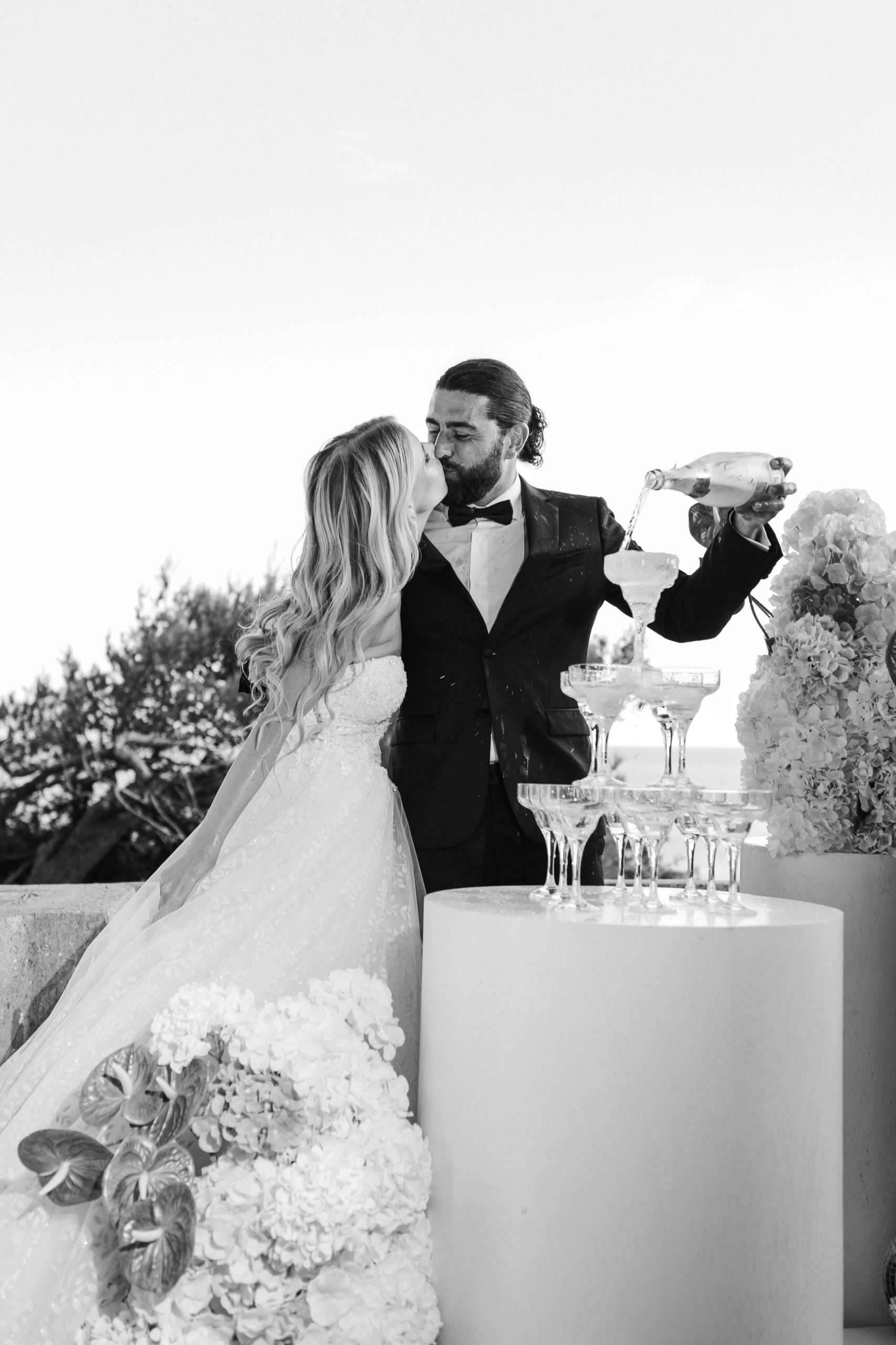 Crno-bijela svadbena fotografija na kojoj mladoženja toči šampanjac u kulu od čaša dok ga nasmiješena mladenka nježno poljubi.