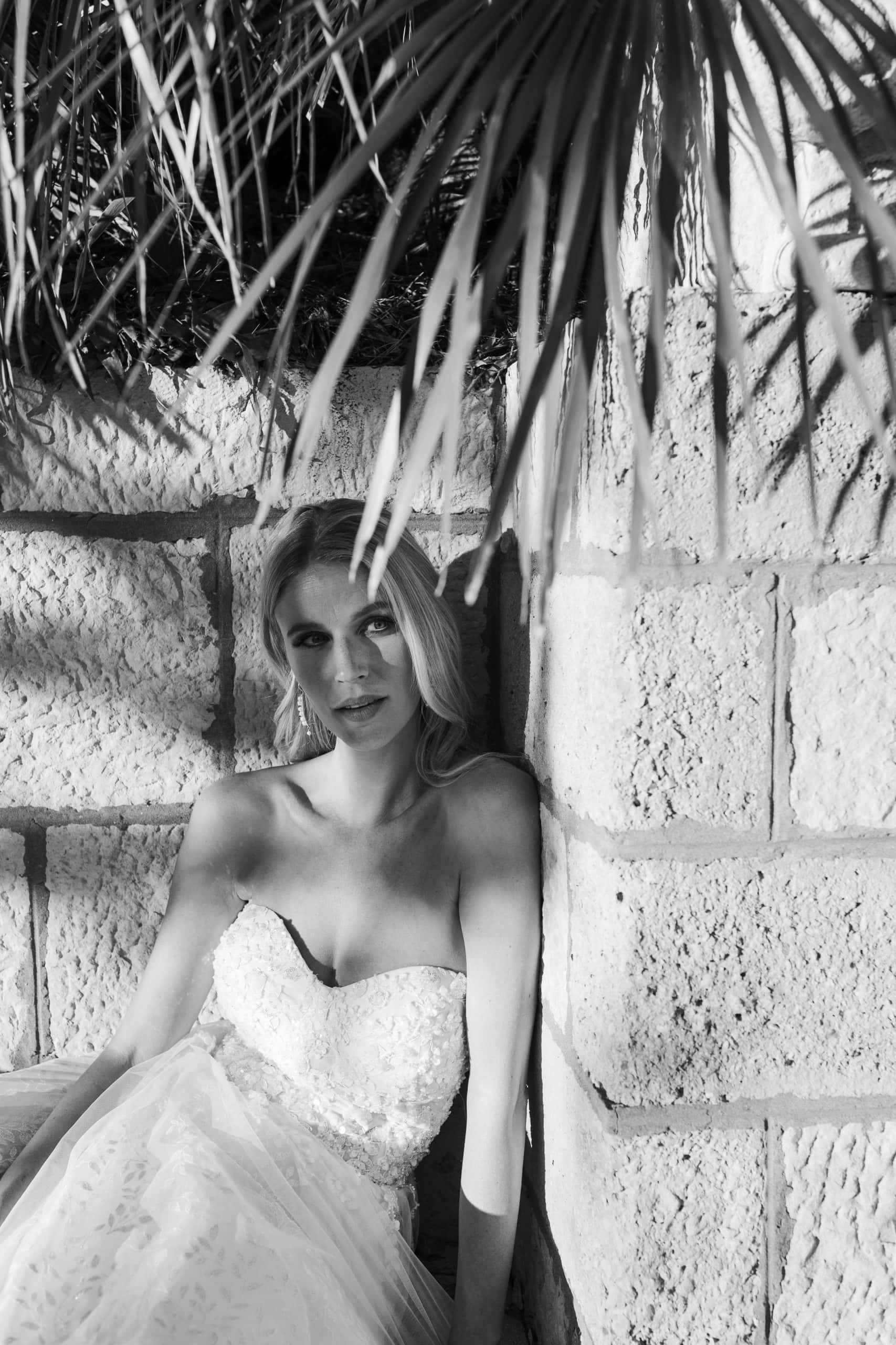 Filmski crno-bijeli editorial portret mladenke u raskošnoj vjenčanici koja sjedi naslonjena na kameni zid ispod palminog lišća, s naglašenom igrom svjetla i sjene.